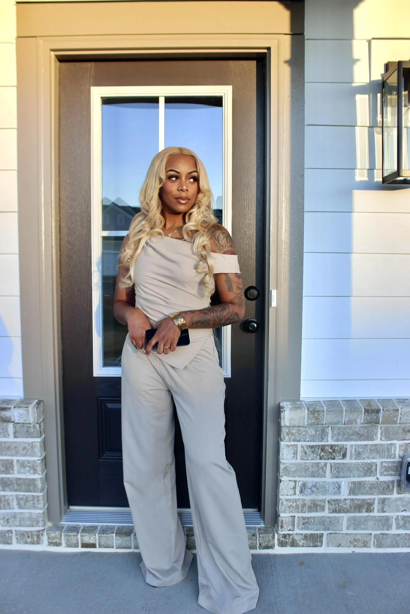 A woman with long blonde hair and tattoos stands in front of a house door, holding a phone, wearing a beige off-shoulder top and matching wide-leg pants, with a reflection of houses and a blue sky in the glass window.