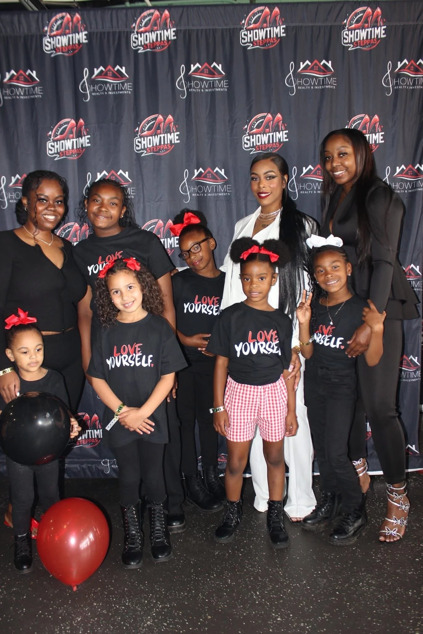 Group of women and children posing in front of a backdrop with 'Showtime Steppas' logos, some children wearing black shirts with 'Love Yourself' printed on them, and balloons on the floor.