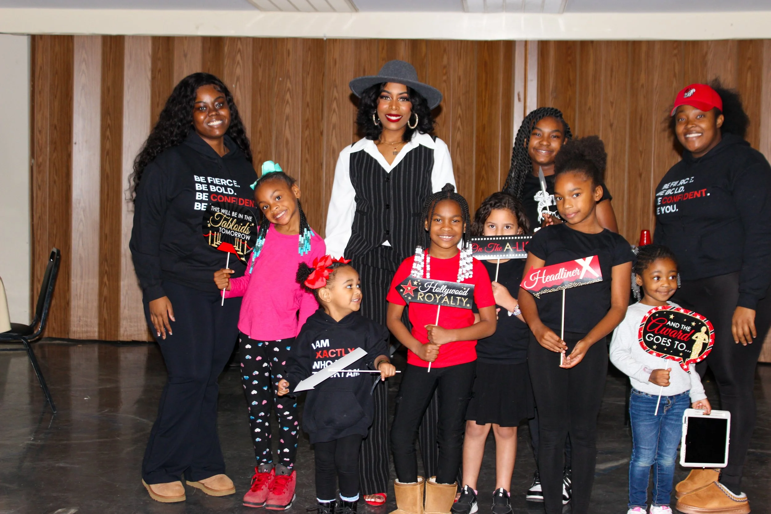 Group of women and children posing together indoors, some holding signs with words like 'Hollywood Loyalty', 'Headliner', and 'And the Award Goes To'. The woman in the center wears a pinstripe suit and a gray hat, while others are dressed casually an