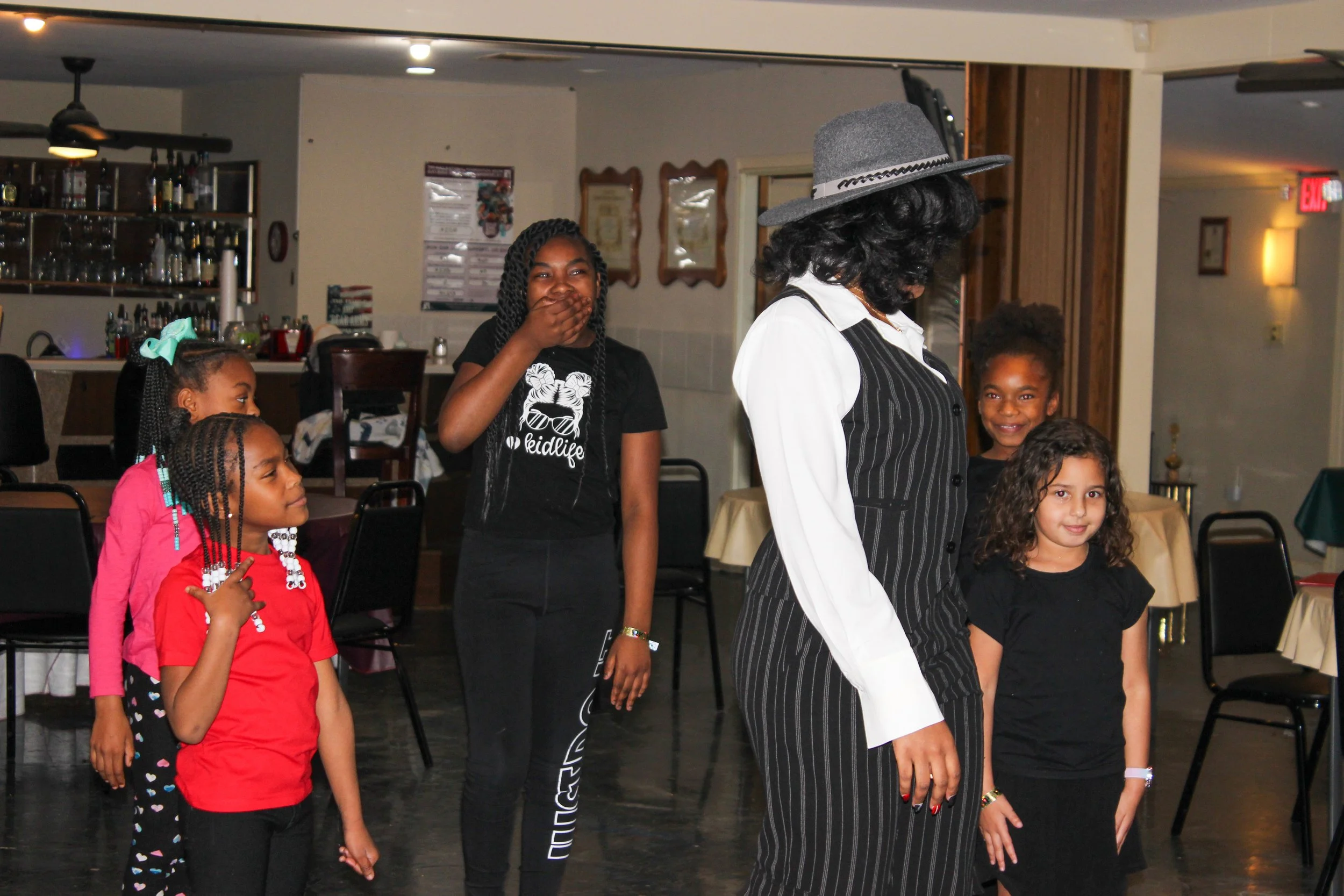 A group of children and a woman standing in a restaurant or dining area, with tables and chairs visible in the background. The woman, dressed in a black pinstripe suit with a white shirt and a gray hat, is looking down while the children around her a