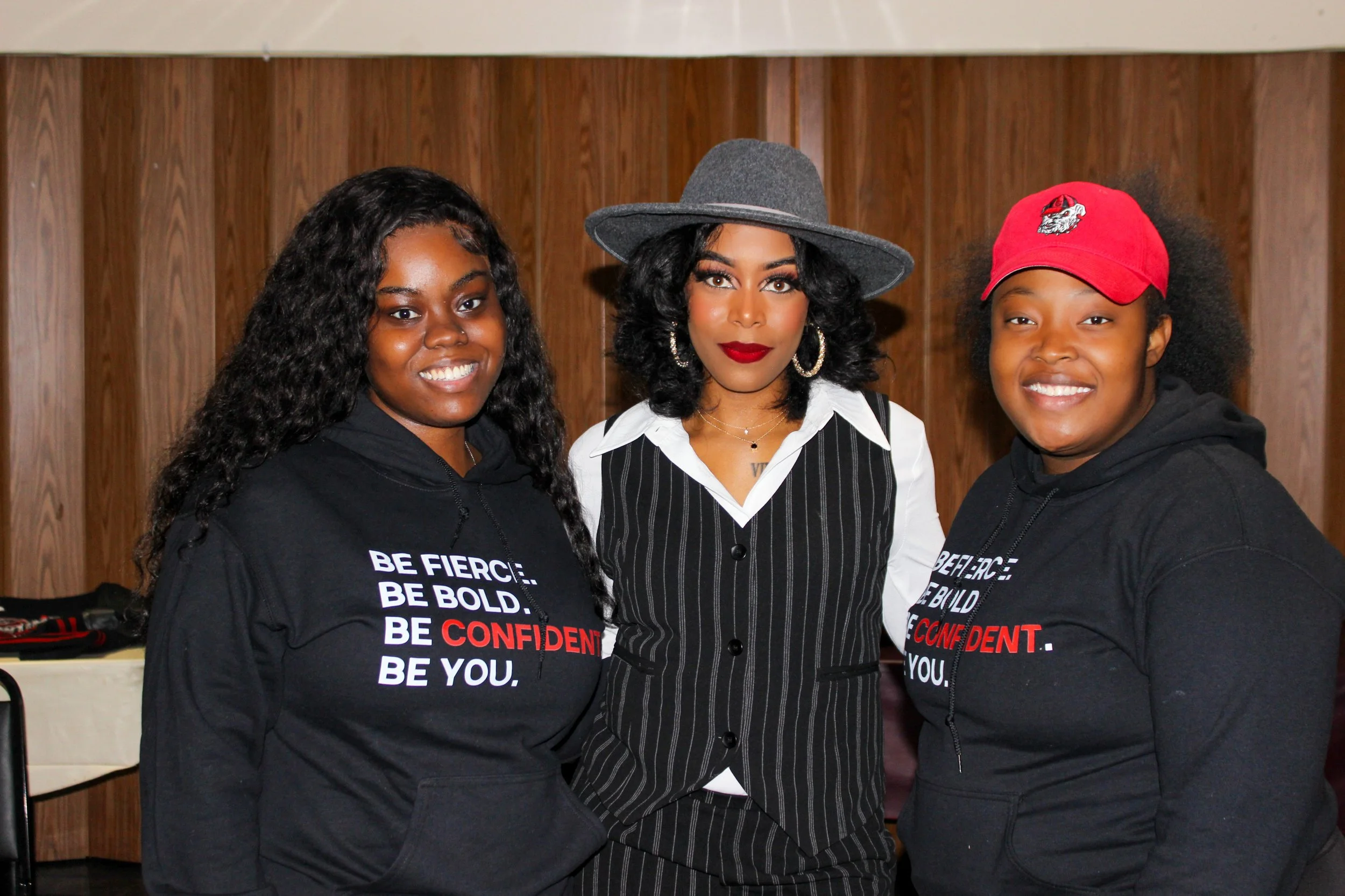 Three women standing together indoors, smiling at the camera. The woman in the middle wears a gray hat, hoop earrings, and a pinstripe vest, while the two women on either side wear black hoodies with motivational slogans.