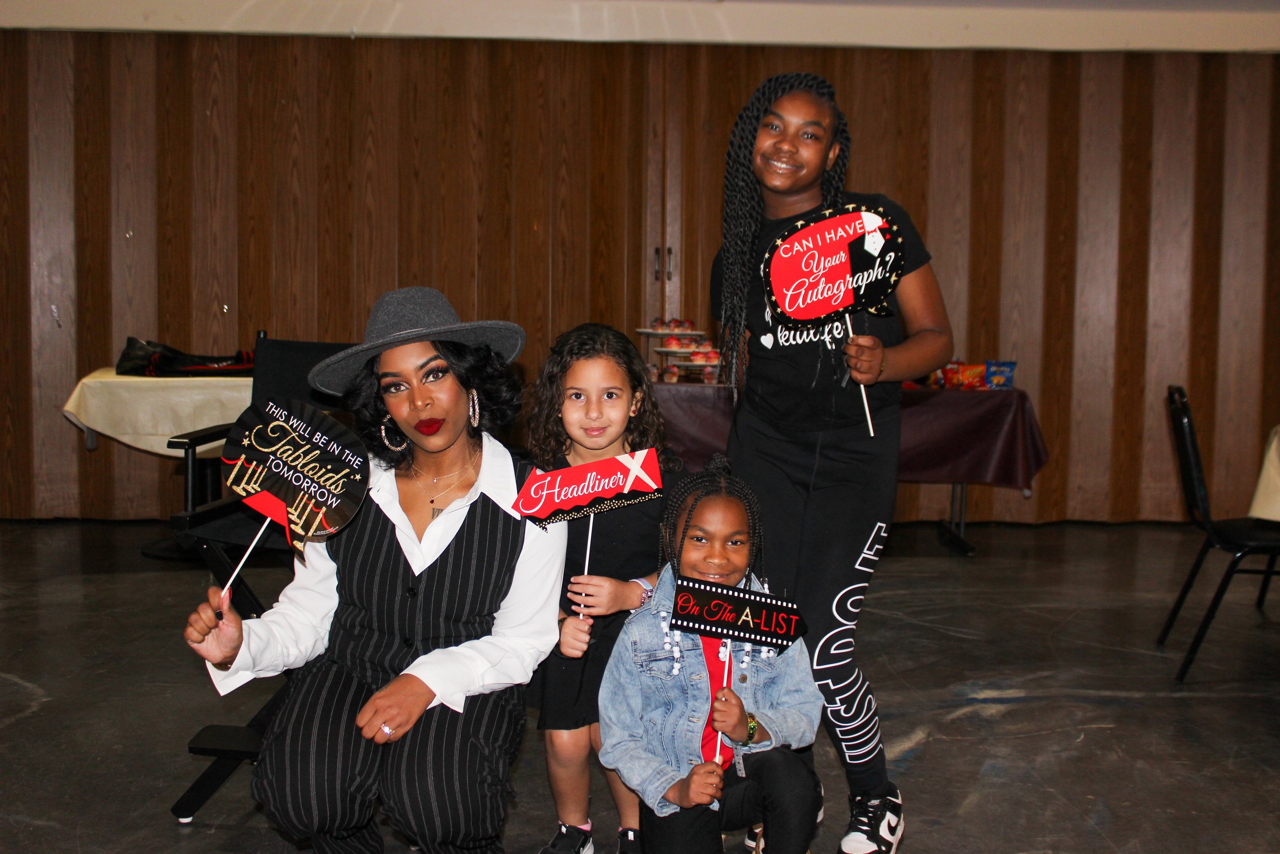 Four women and girls posing for a photo at a celebration, holding signs that say 'This will be the baddest (sic) TALBIDS tomorrow,' 'Headliner,' 'Can I have your autograph?,' and 'On the A LIST.' They are smiling and dressed stylishly, with a wooden 