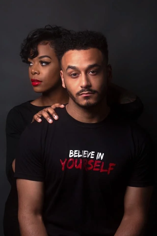 A man and woman pose together against a dark background, with the woman standing behind and resting her hand on the man's shoulder. The man wears a black T-shirt that says 'Believe in Yourself' in red and white lettering. The woman has short curly ha