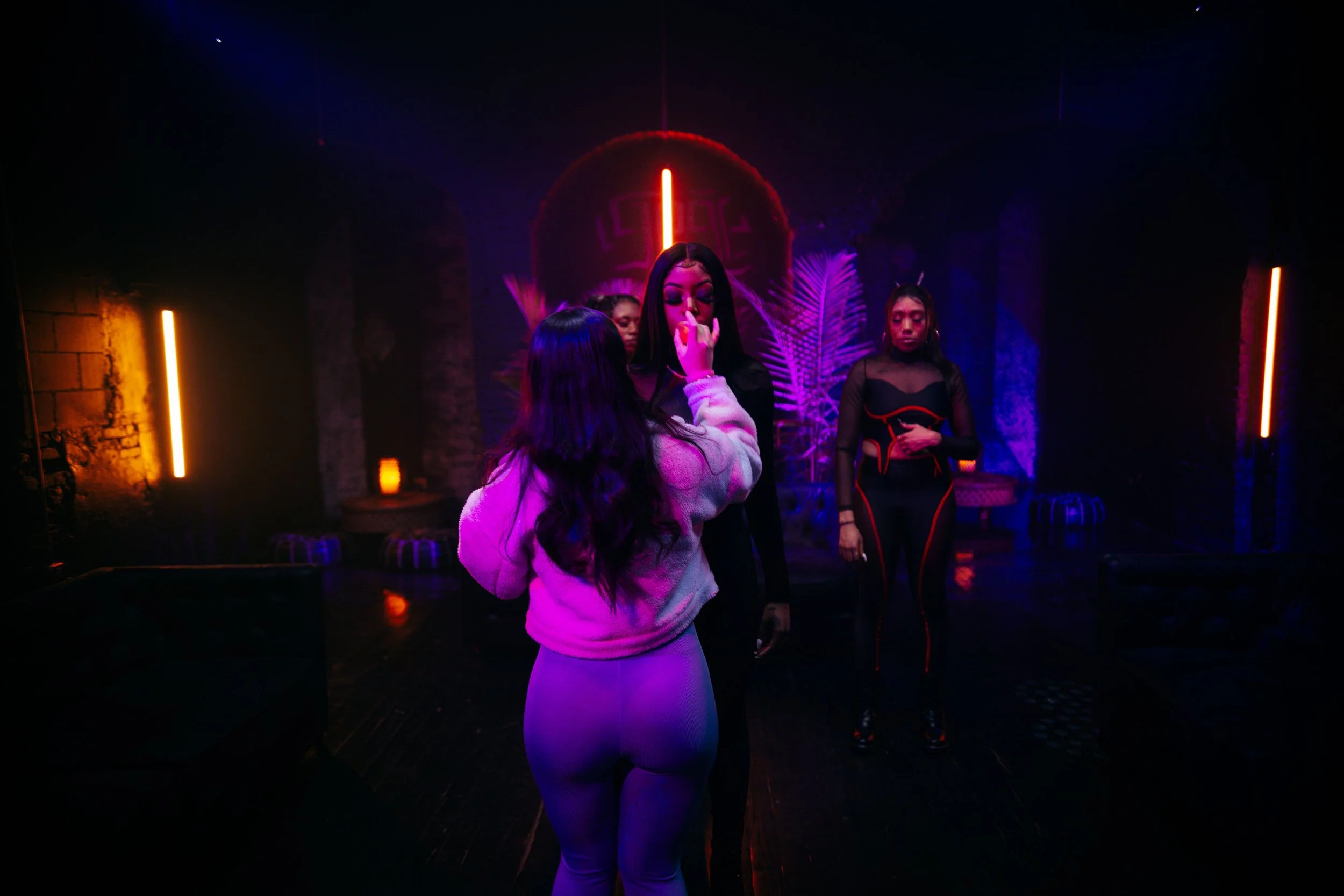 Three women in a dimly lit room with vibrant neon lights; one woman is applying makeup or makeup touch-up to another, while a third woman stands nearby observing, with tropical plants and minimal furniture in the background.