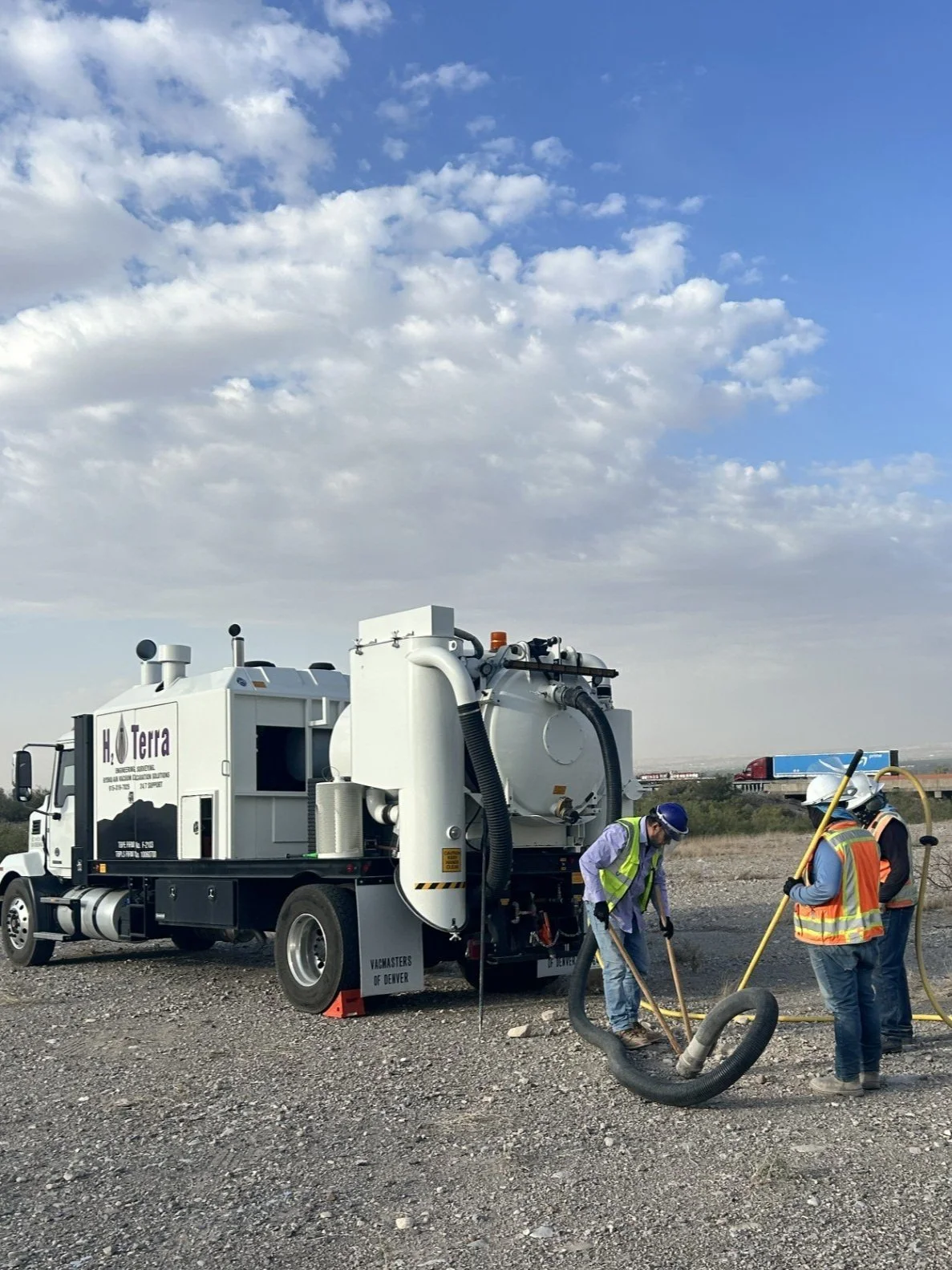 Three workers in safety vests and helmets operate a vacuum truck on a gravel lot, with a blue sky and scattered clouds overhead.