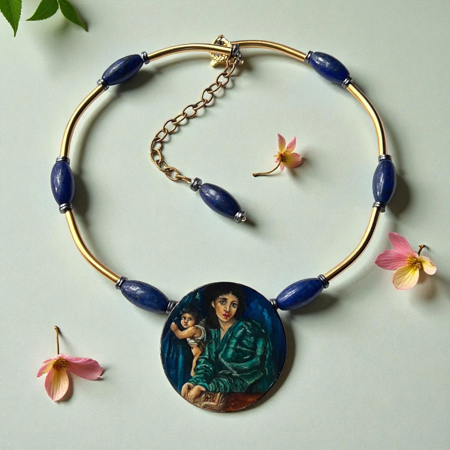 A necklace with large blue beads and a round pendant featuring a painted portrait of a woman holding a child, laid on a light green surface with scattered pink flowers.