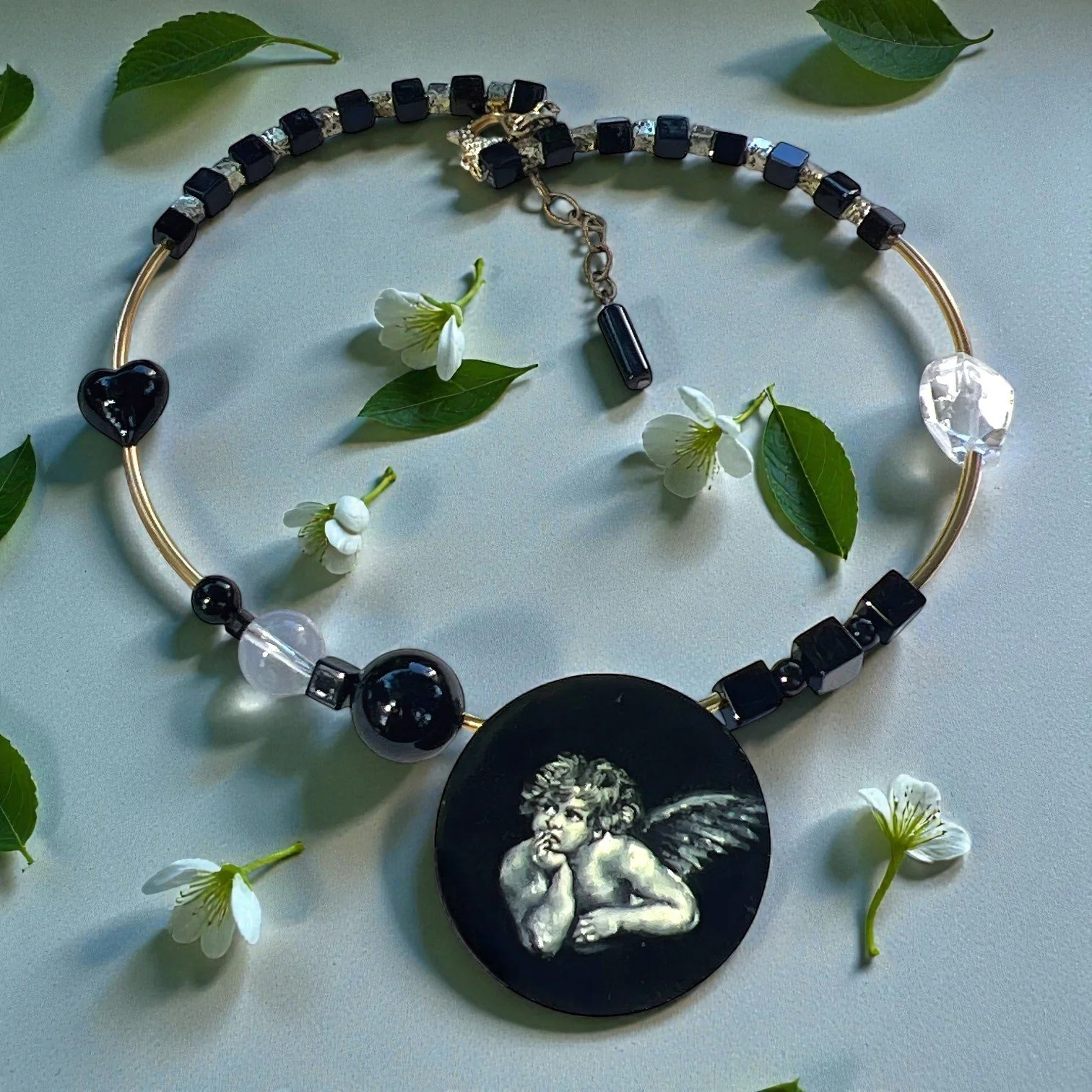 A black and gold beaded necklace with a large round pendant featuring a cherub, surrounded by white flowers and green leaves.