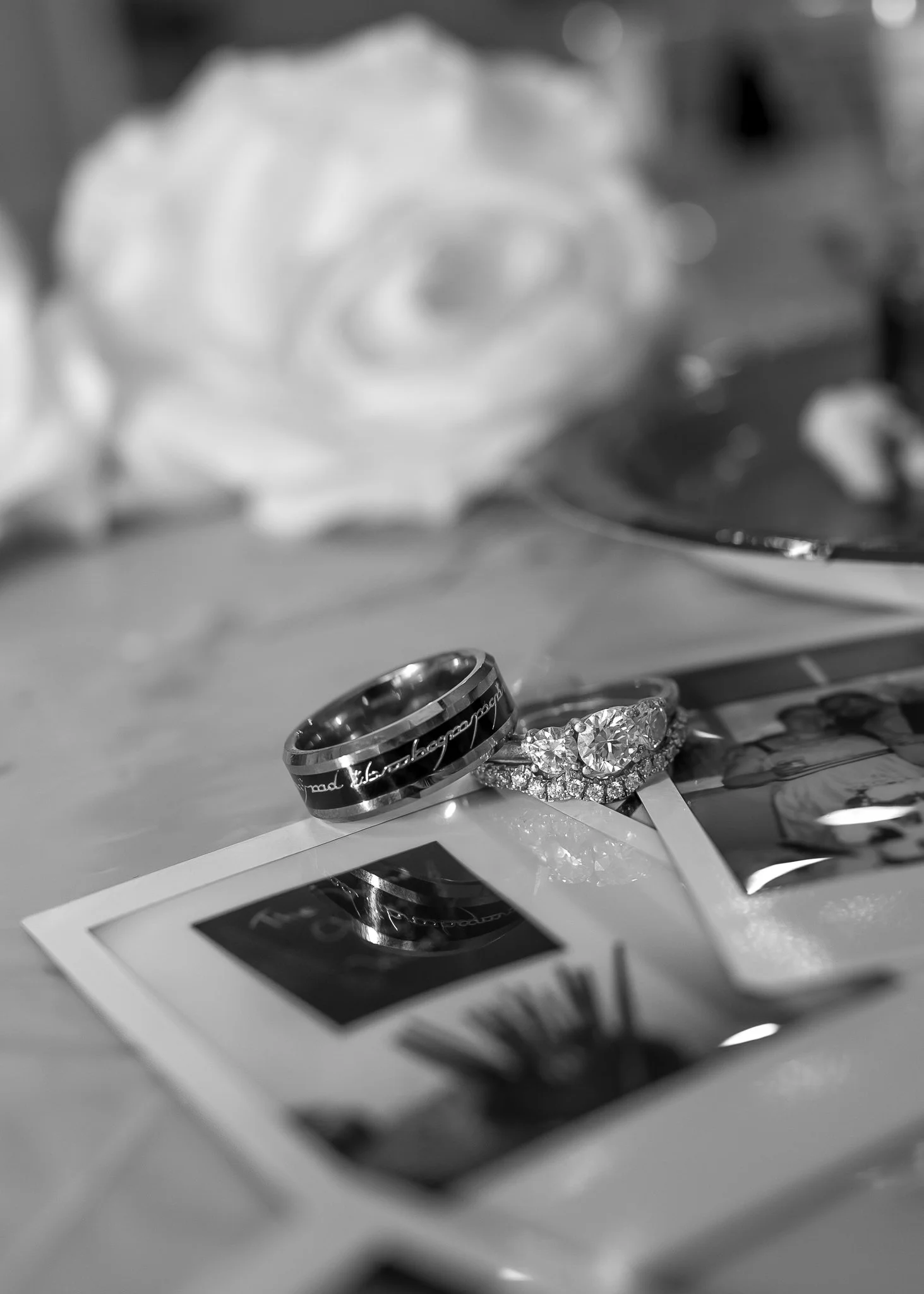 Close-up of wedding rings, one with a diamond, placed on a photograph with a white rose in the blurred background.