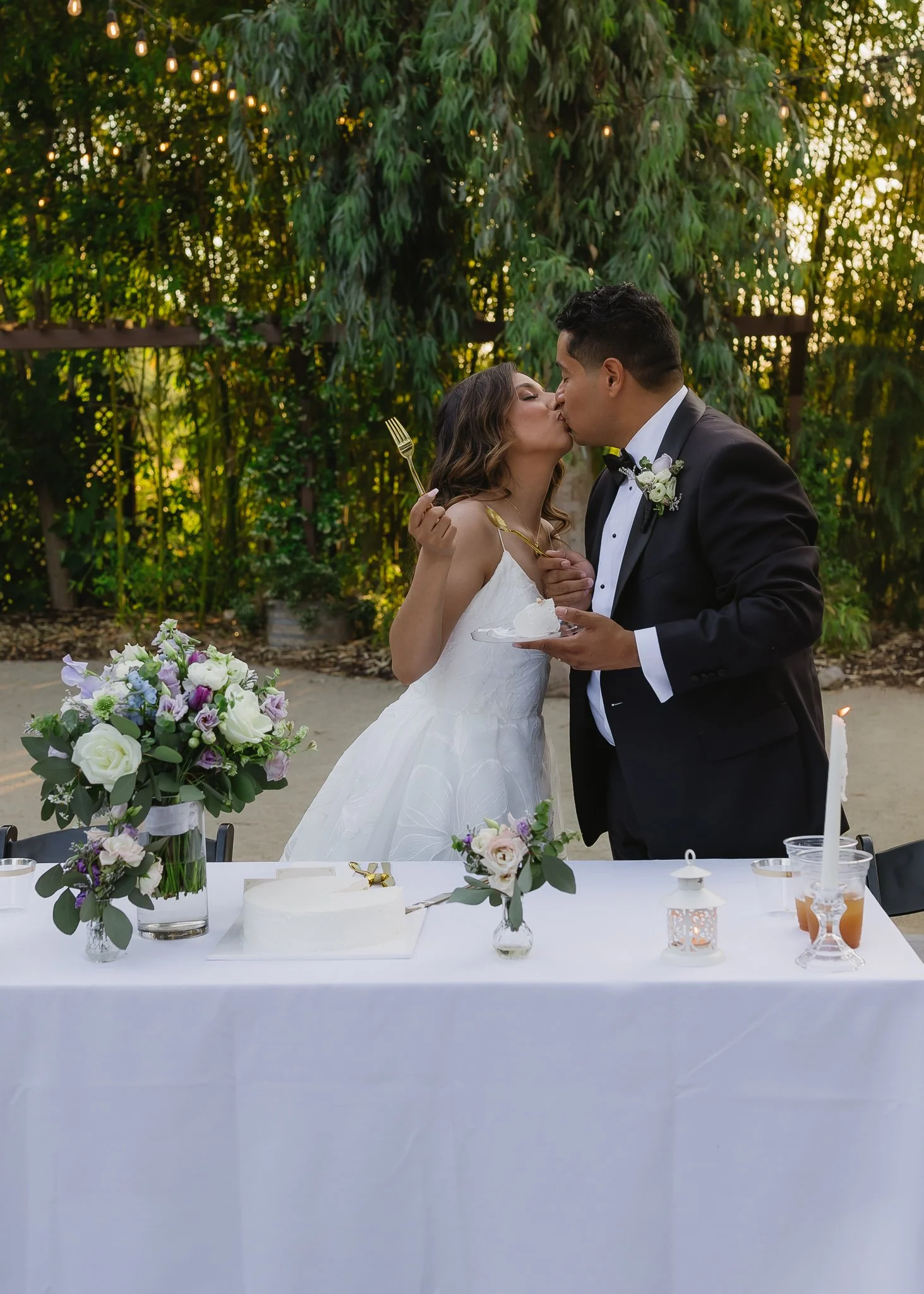 A bride and groom kiss at their wedding reception, standing behind a table with floral arrangements, a cake, and candles, outdoors with greenery in the background.