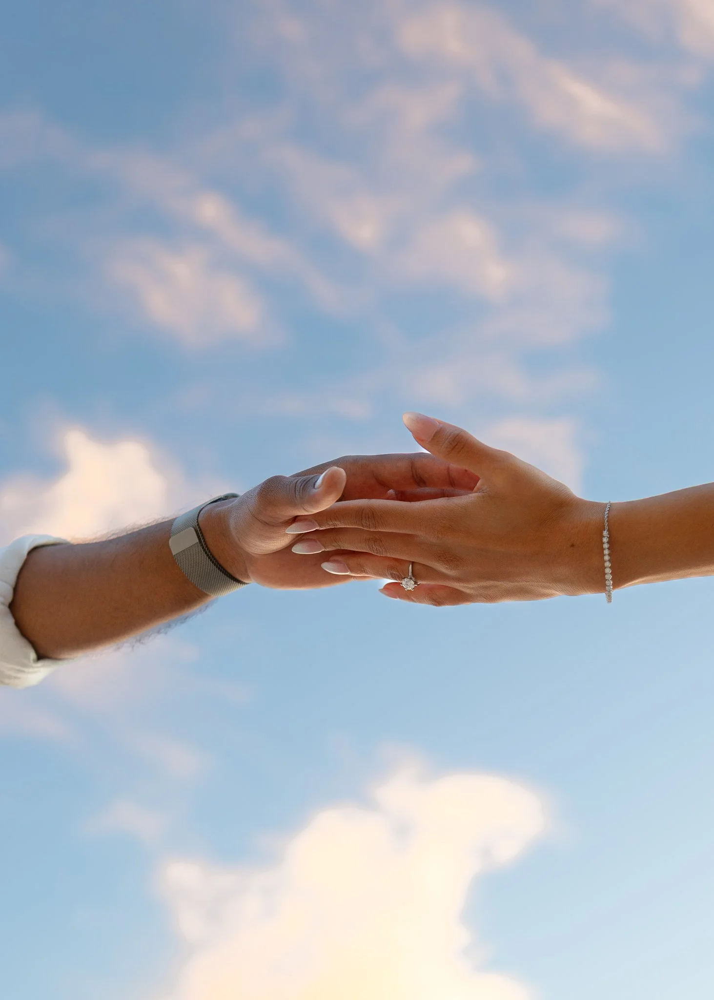 Two hands, one male and one female, reach towards each other with fingers touching against a blue sky with clouds.
