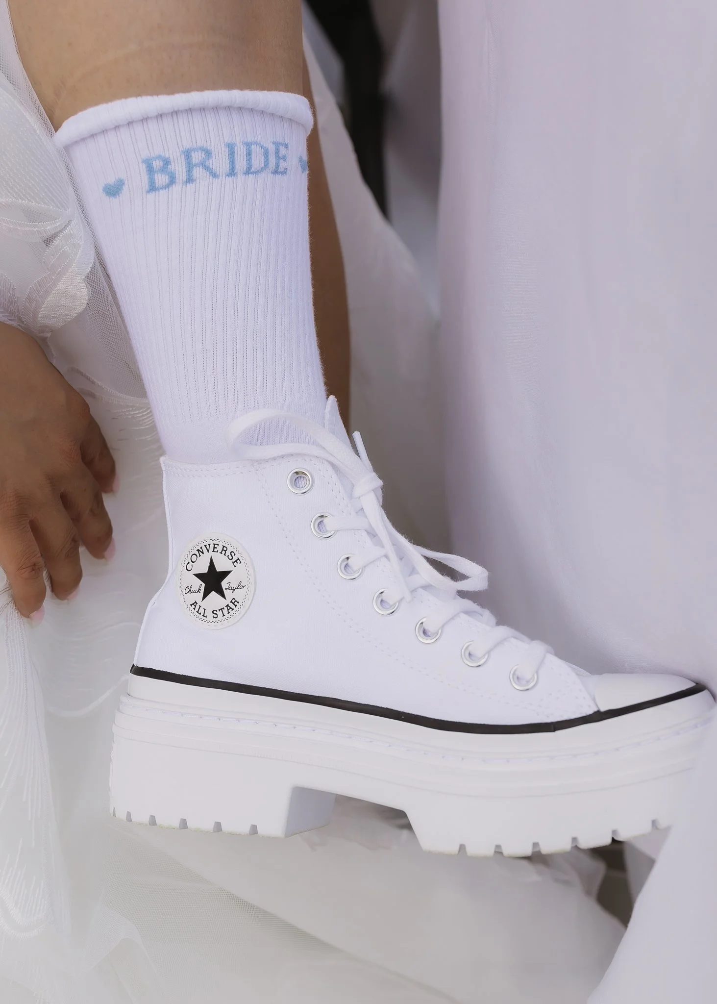 A white high-top Converse sneaker on a person's foot, with a thick platform sole and a 'Bride' sock in light blue with small hearts, against a white background.