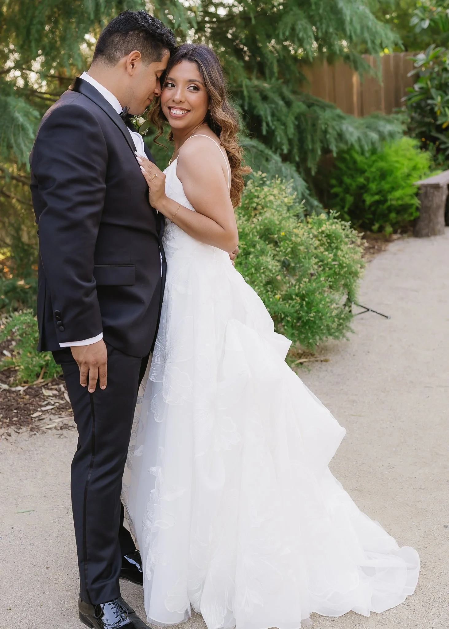 A newlywed couple in wedding attire standing close together outdoors in a lush green garden, smiling and sharing an intimate moment.