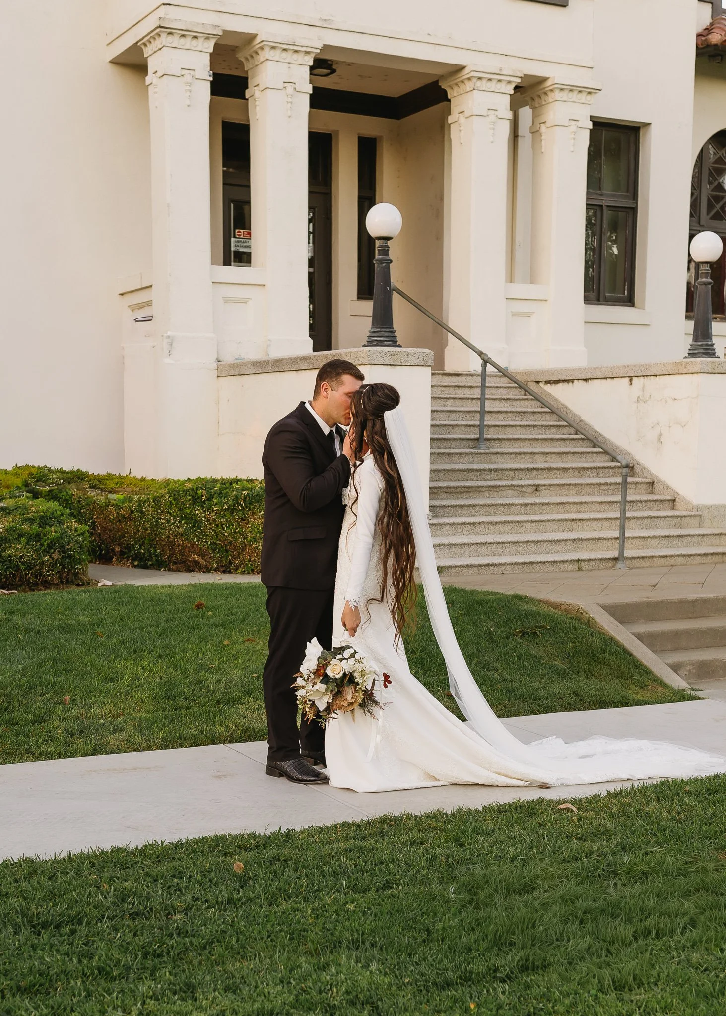A bride and groom share a kiss outside a building with white columns and stairs. The bride is holding a bouquet of flowers and wearing a long white wedding dress with a veil. The groom is dressed in a black tuxedo.