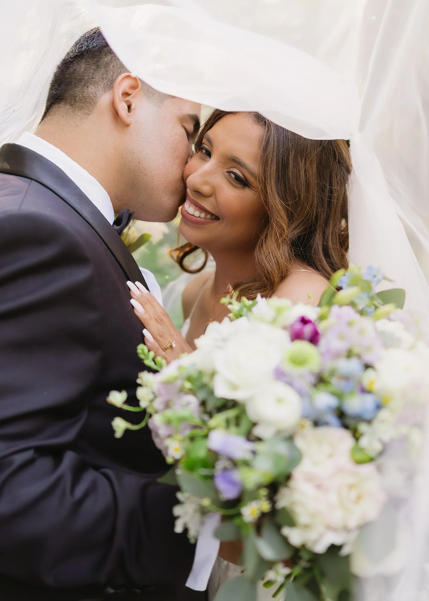 A newlywed couple sharing a kiss under a wedding veil, with the bride smiling and holding a bouquet of flowers.