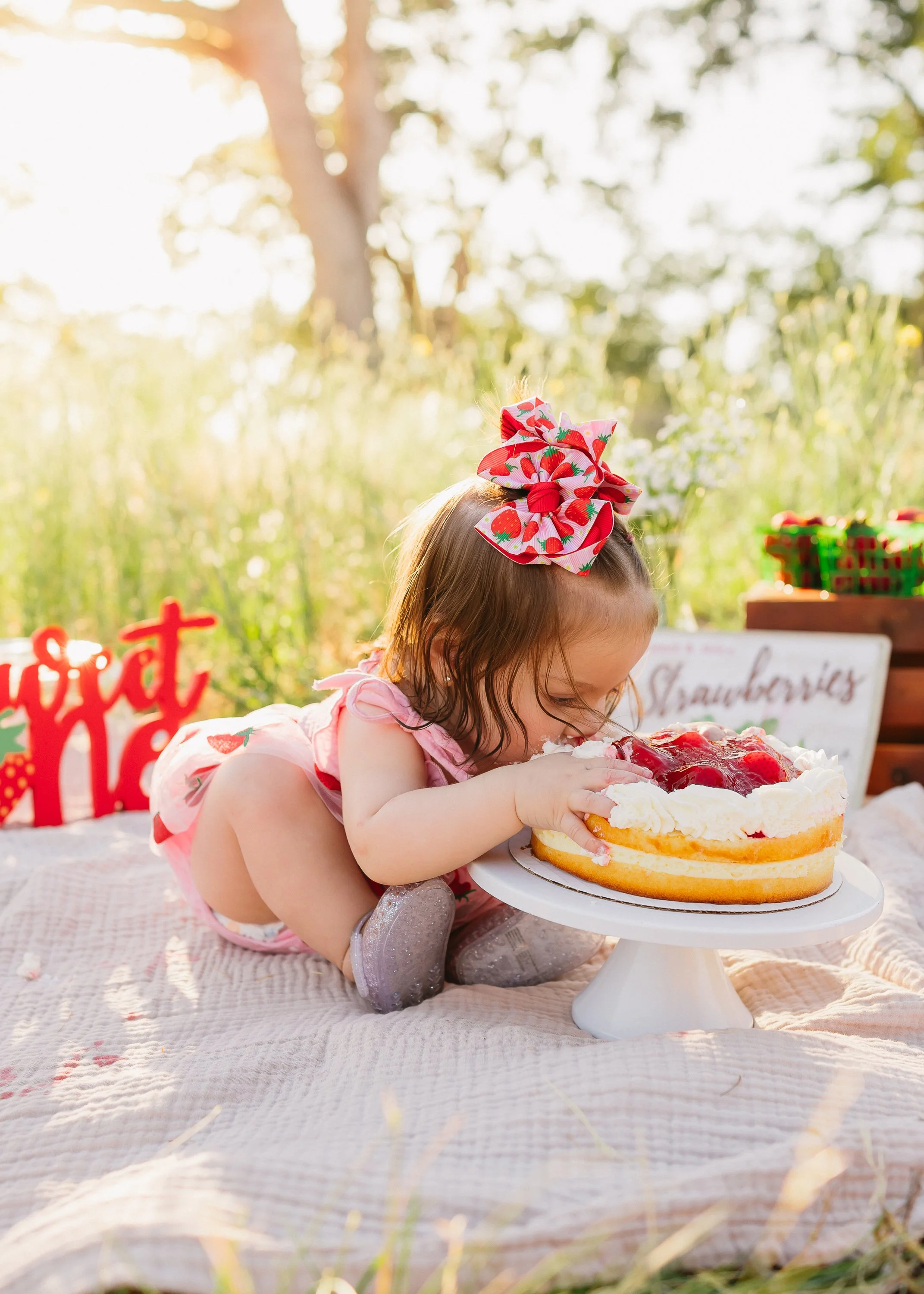 A young girl with a large red and white strawberry-themed hair bow is leaning over a strawberry shortcake on a cake stand outdoors during daytime. The scene is set in a grassy field with trees and a picnic setup in the background.