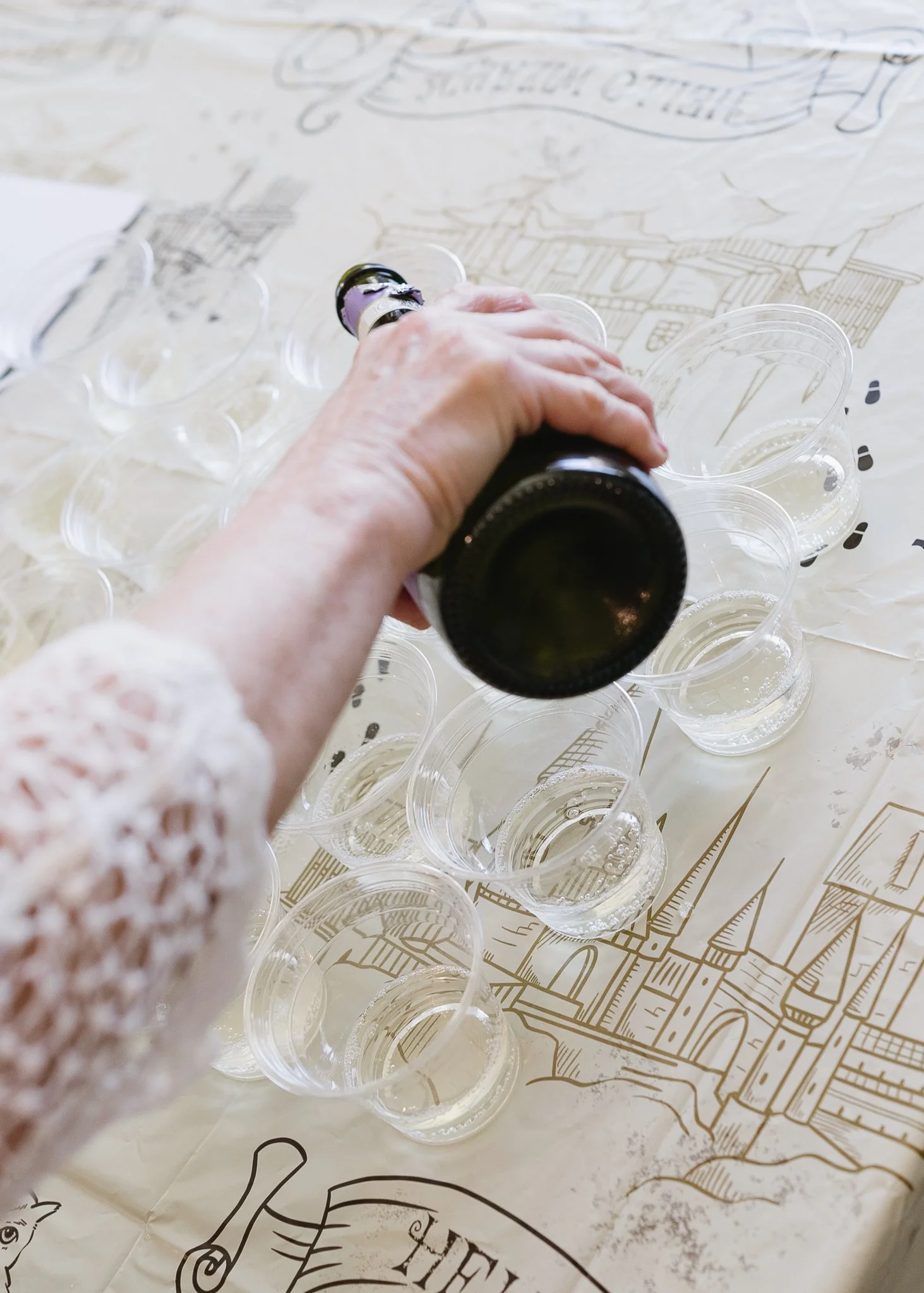 A person wearing a lacy white sleeve holding a dark bottle, pouring a clear liquid into small, clear plastic cups arranged on a beige paper with a castle and text design.