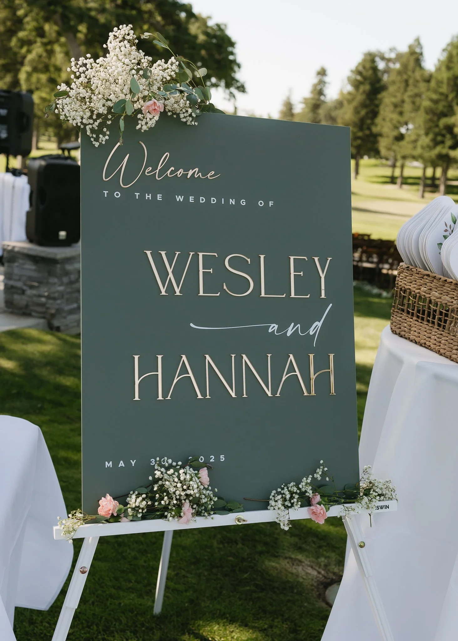 Wedding welcome sign with floral decorations, announcing the wedding of Wesley and Hannah on May 30, 2025, outdoors on a grassy area with trees in the background.