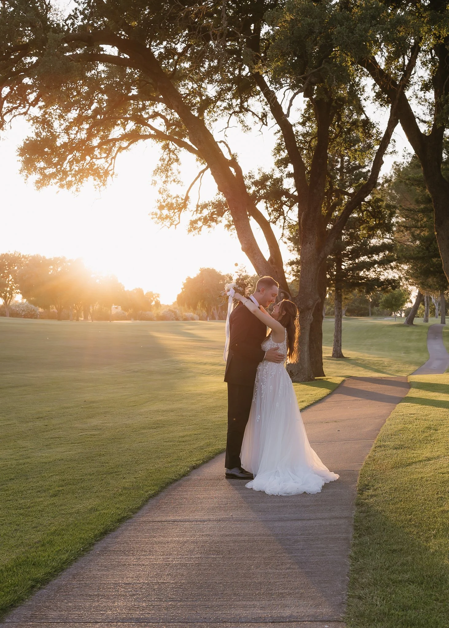 A bride and groom embrace on a park pathway at sunset, surrounded by tall trees with sunlight filtering through the leaves.