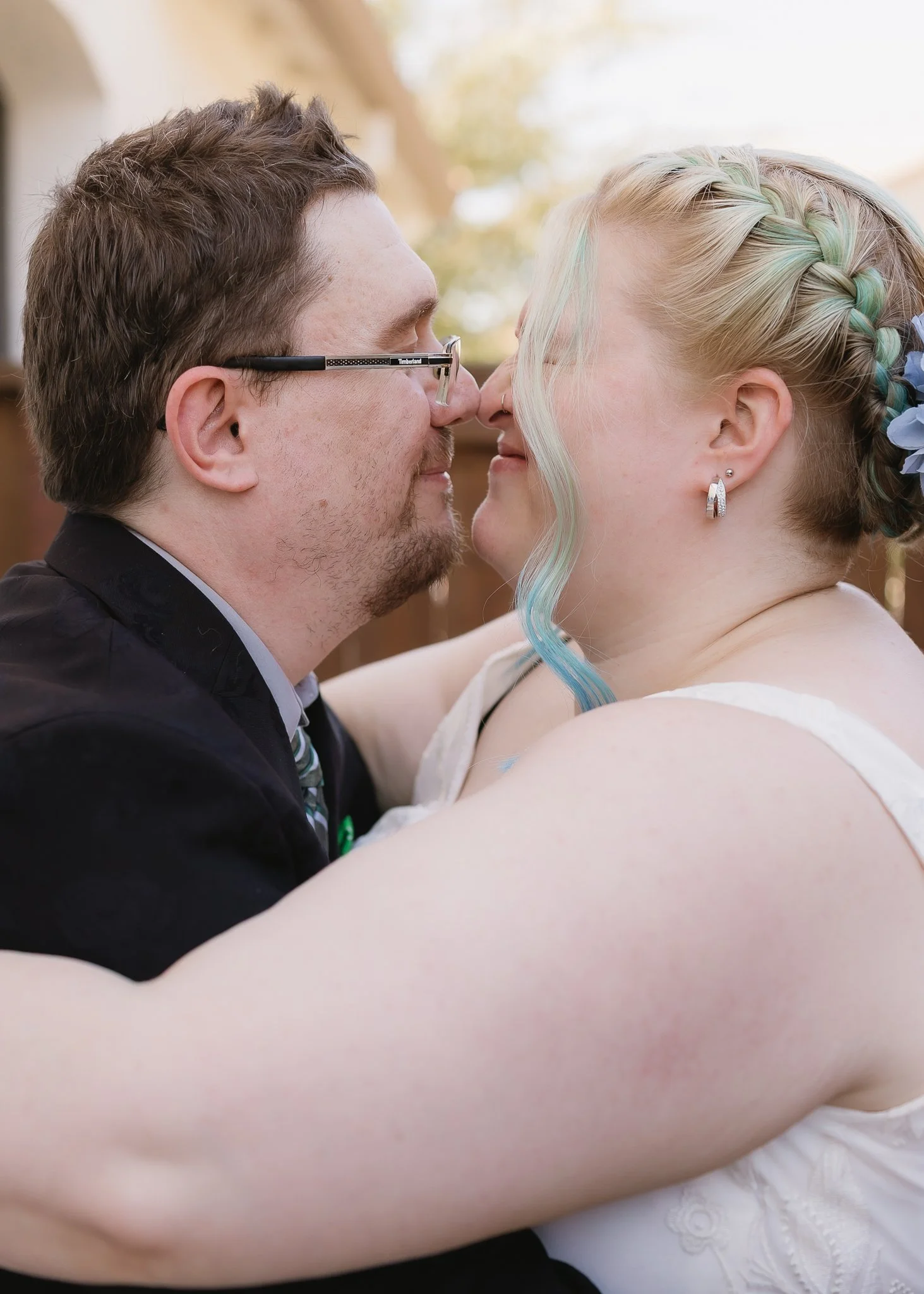 A close-up of a couple touching noses, embracing outdoors during daylight, with the man wearing glasses and the woman with braided hair and pastel-colored highlights.