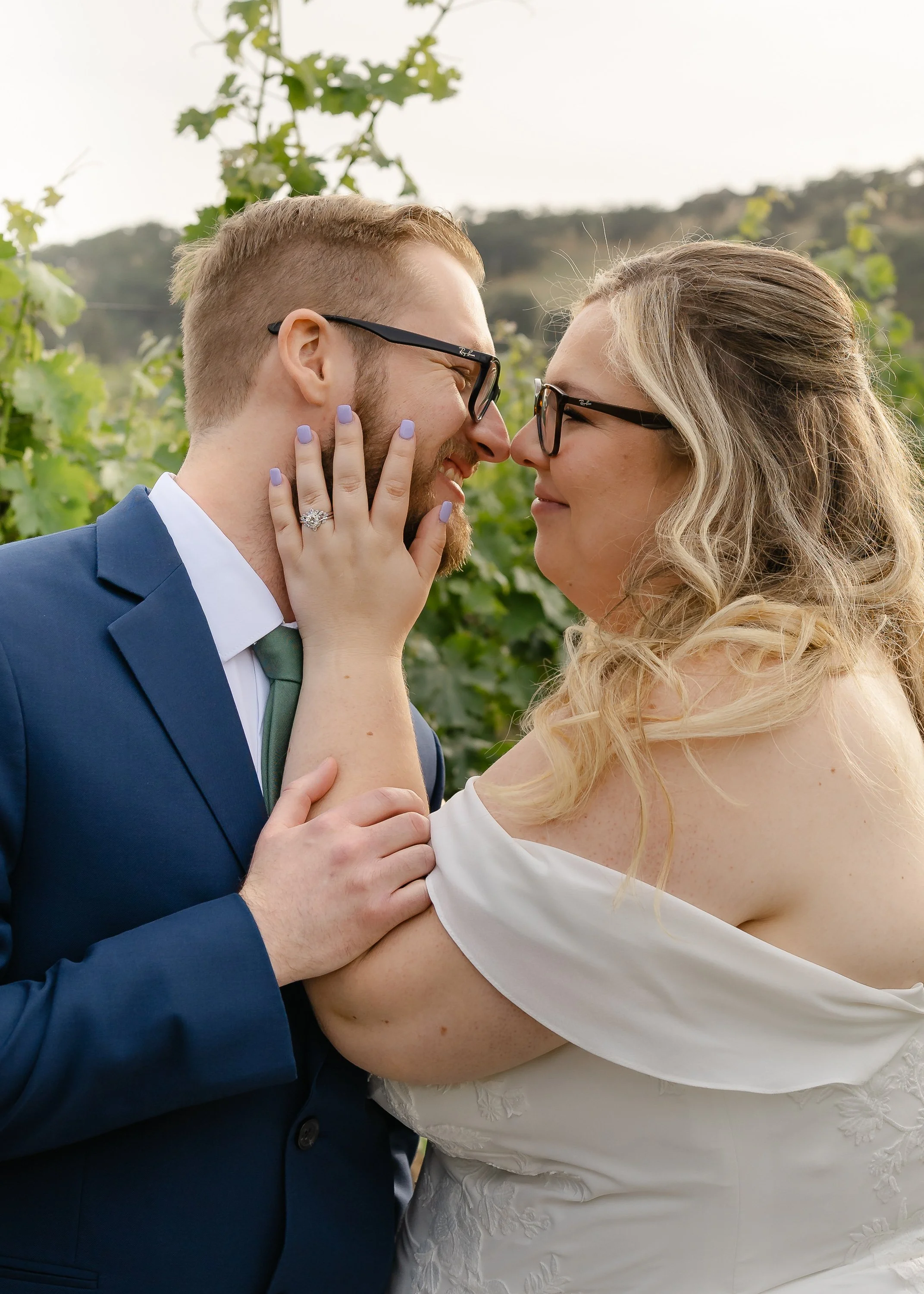 A couple in wedding attire, the man in a blue suit and the woman in a white dress, face each other closely outdoors with greenery and hills in the background, smiling with their foreheads and noses touching.
