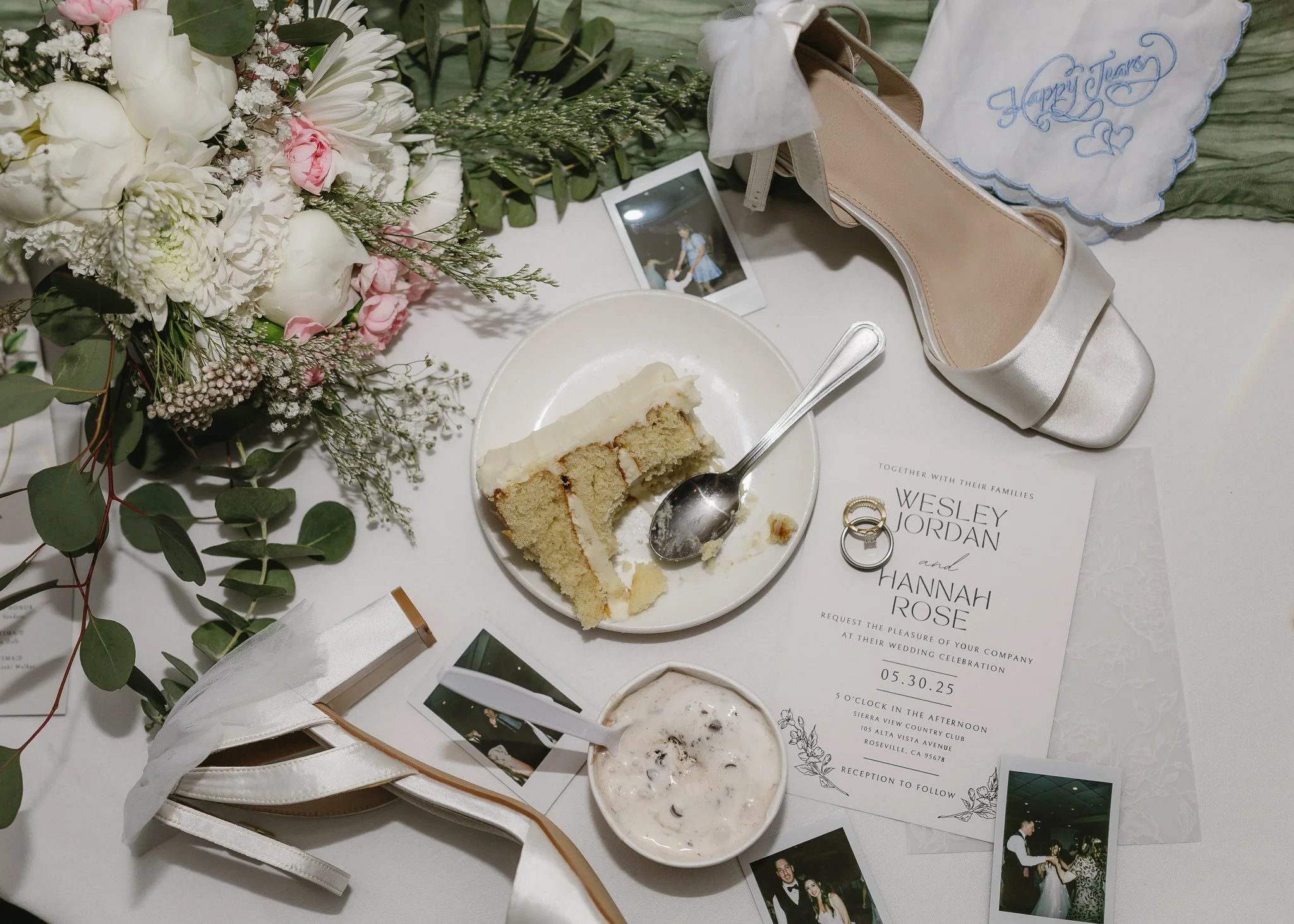 A table at a wedding reception with a flower arrangement, a wedding invitation, a slice of cake on a plate, a cup of iced coffee, a cream-colored satin shoe with a bow, two Polaroid photos, and a white cloth with 'Happy Tears' embroidered in blue.