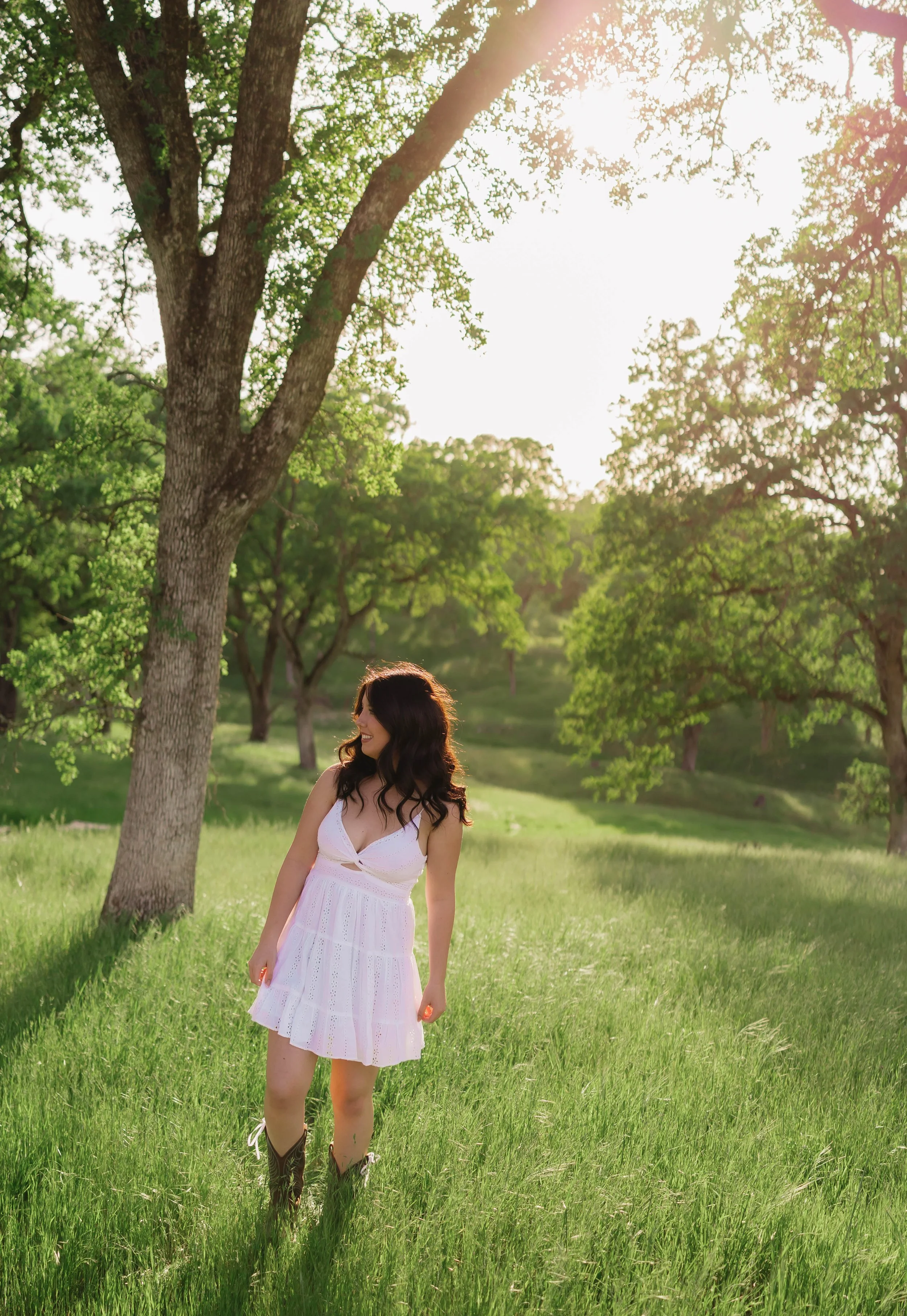 A woman in a white dress standing in a green, sunlit meadow surrounded by trees.