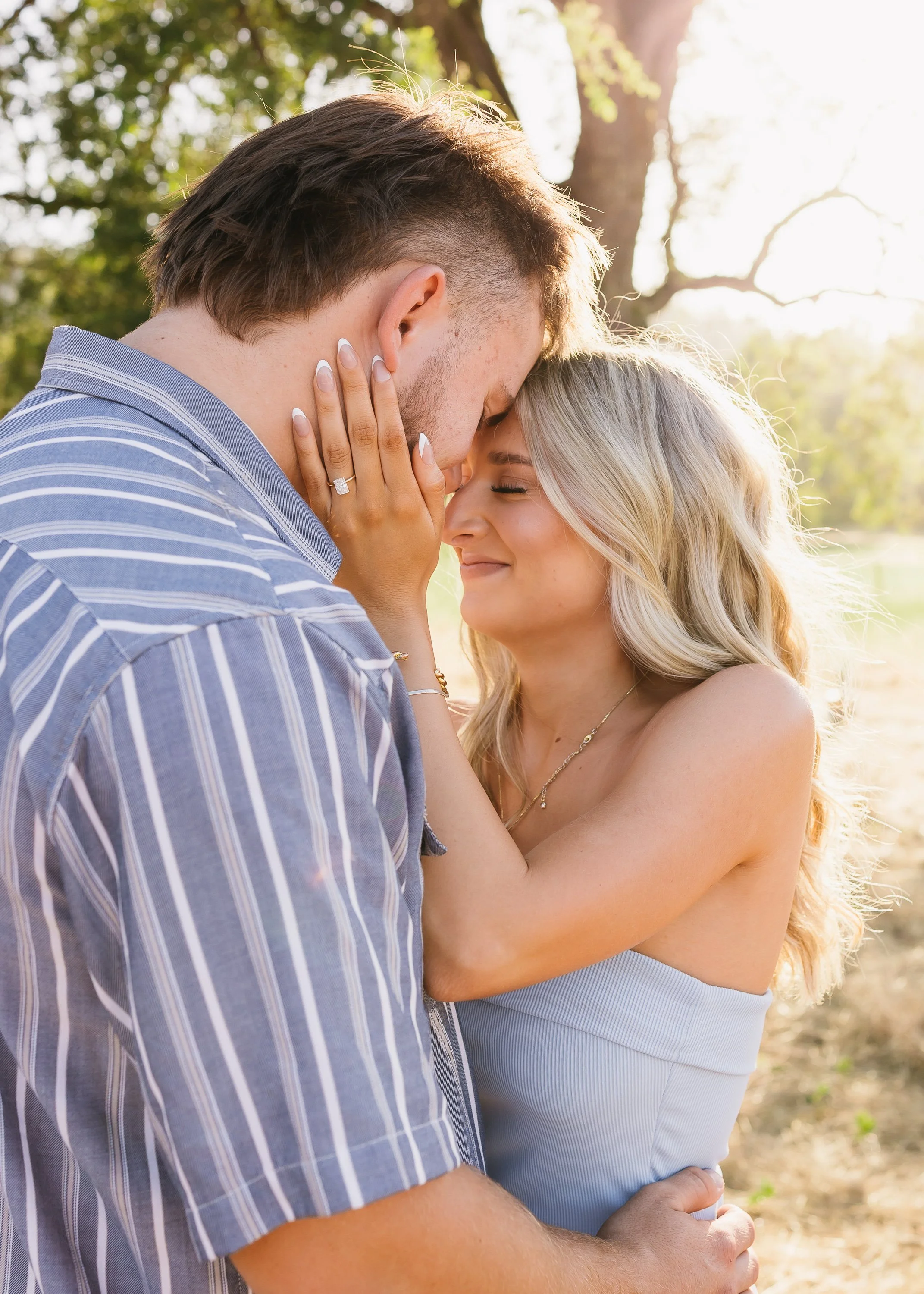 A couple shares a romantic moment outdoors, with the man touching foreheads and the woman smiling with eyes closed, sunlight filtering through trees in the background.