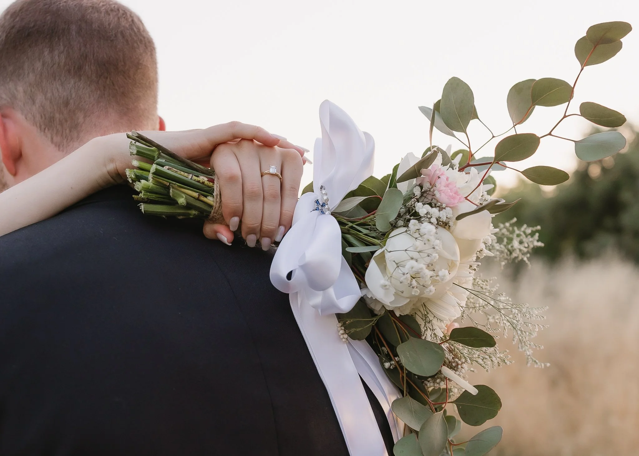 A bride holding a bridal bouquet with white and pink flowers and greenery on a groom's shoulder, with a background of blurred trees and sky.