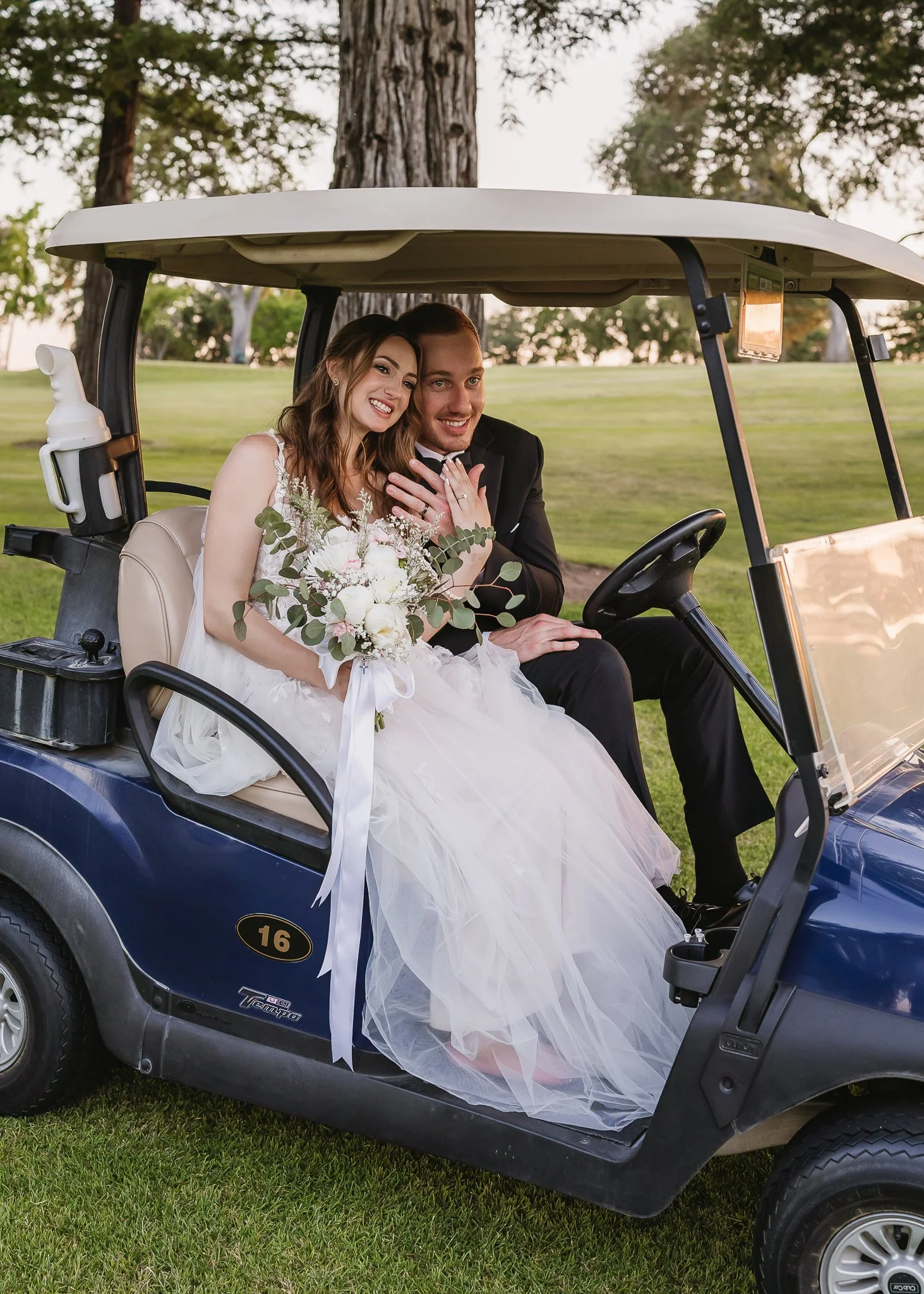 A newlywed couple sitting inside a golf cart on a golf course. The bride is holding a bouquet, and the groom is dressed in a black tuxedo. They are smiling and looking happy.