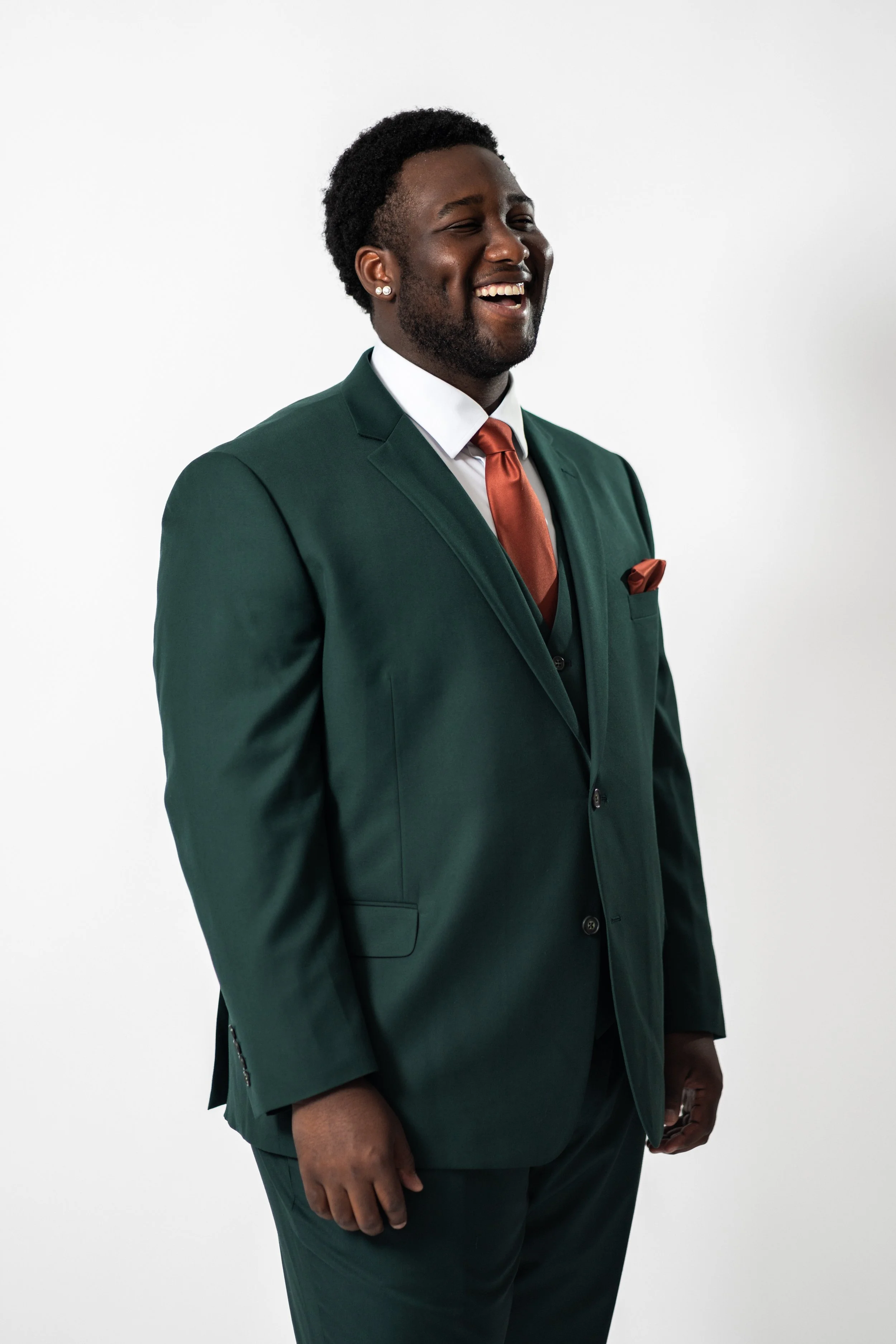 A smiling man in a dark green suit with a white shirt, rust-colored tie, and matching pocket square stands against a plain white background.