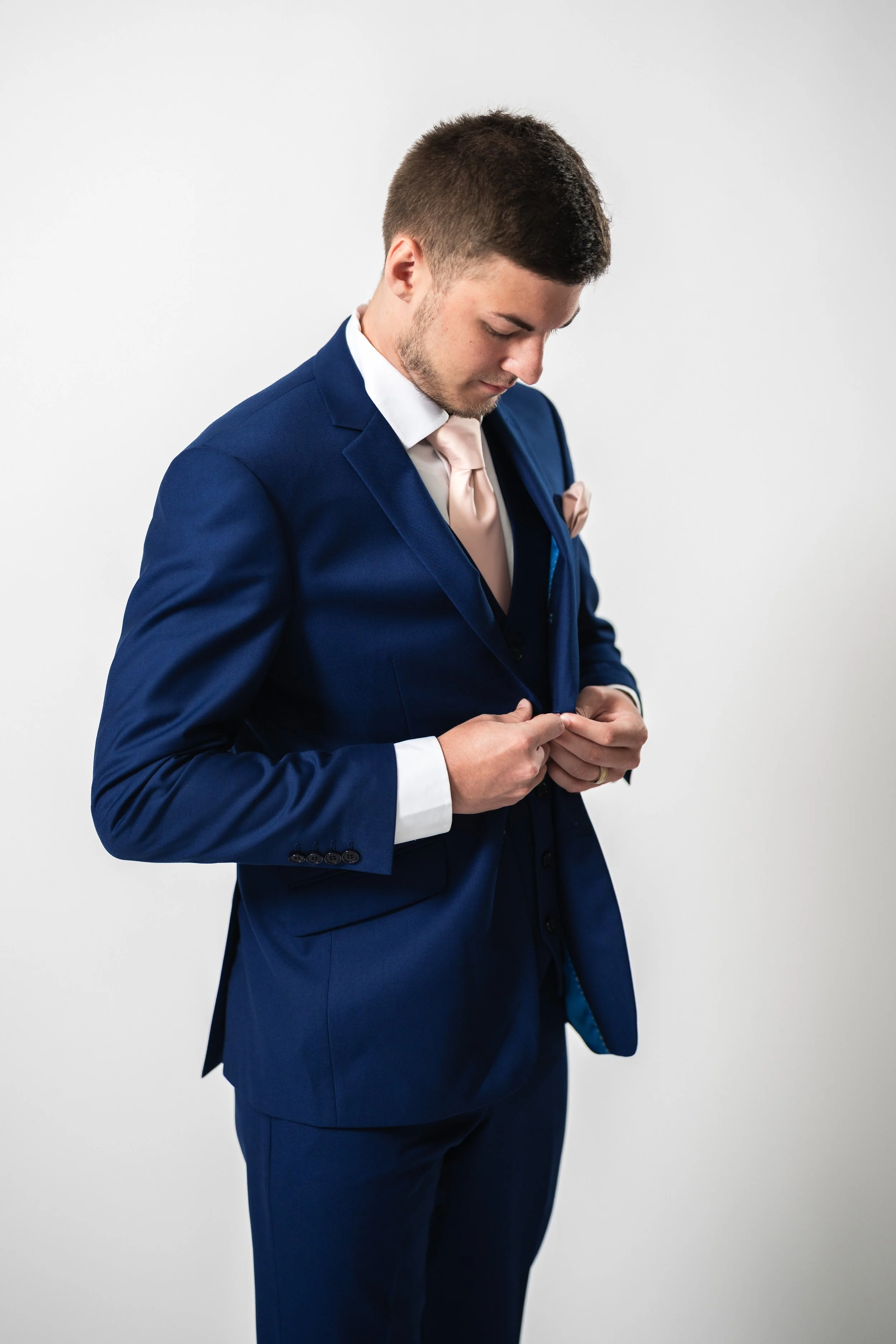 Young man in a blue suit buttoning his jacket, standing against a plain white background.