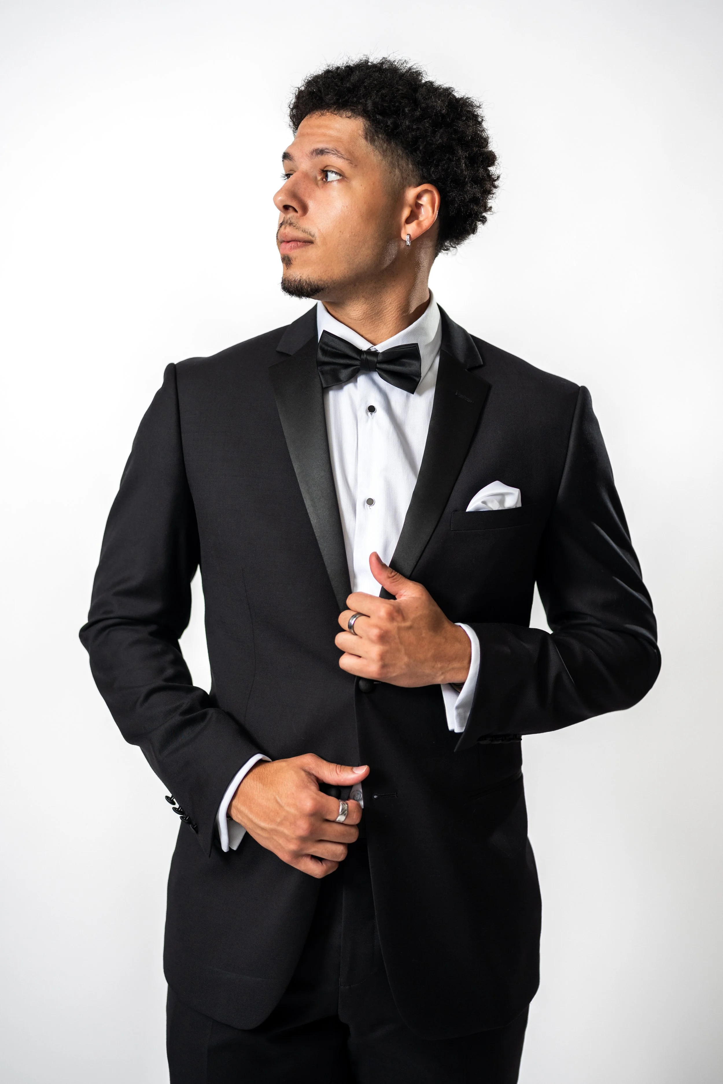 A man dressed in a black tuxedo with a white shirt and black bow tie, standing against a plain white background.