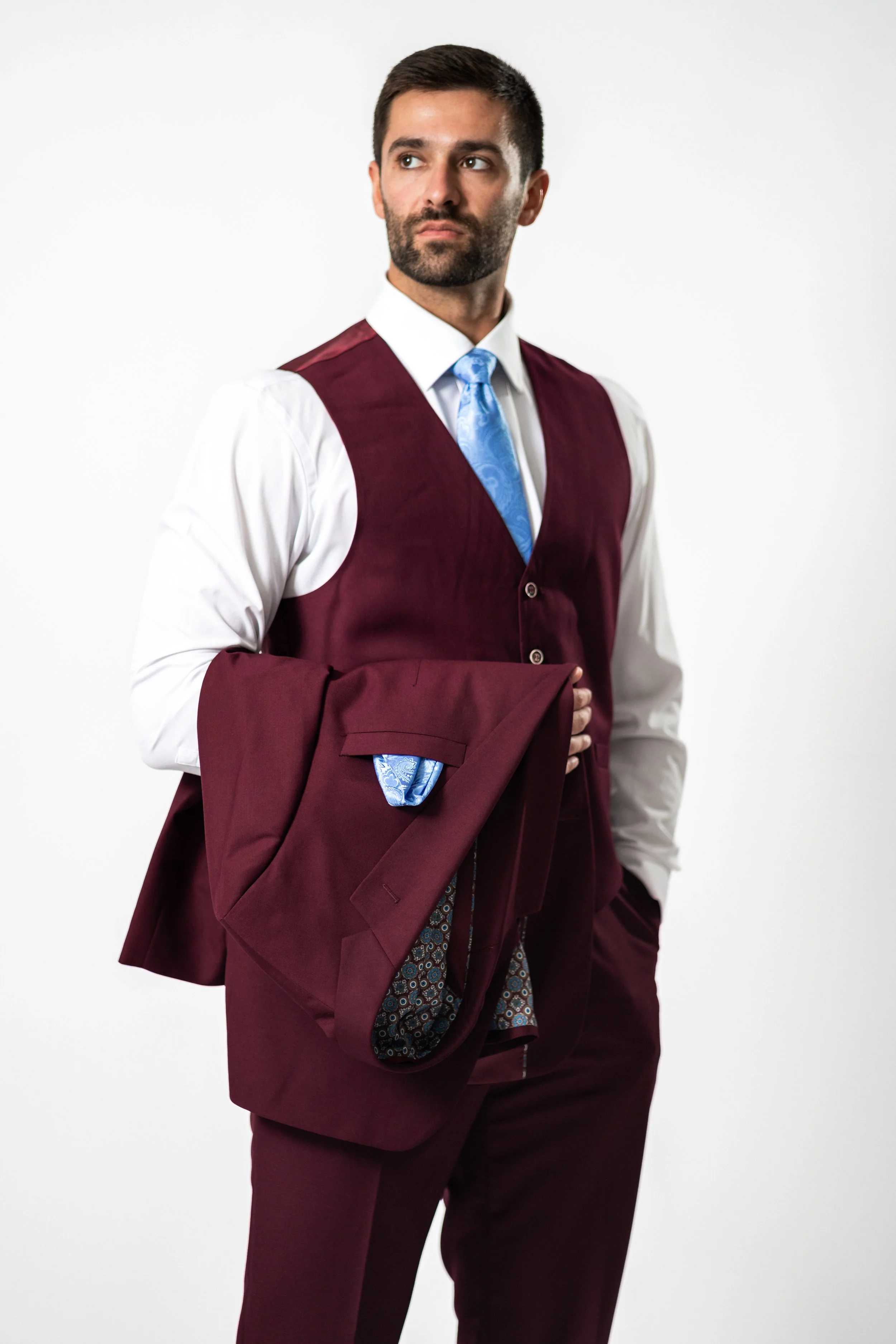 A man in a burgundy suit with a light blue tie, holding a matching burgundy blazer with patterned lining, against a plain white background.