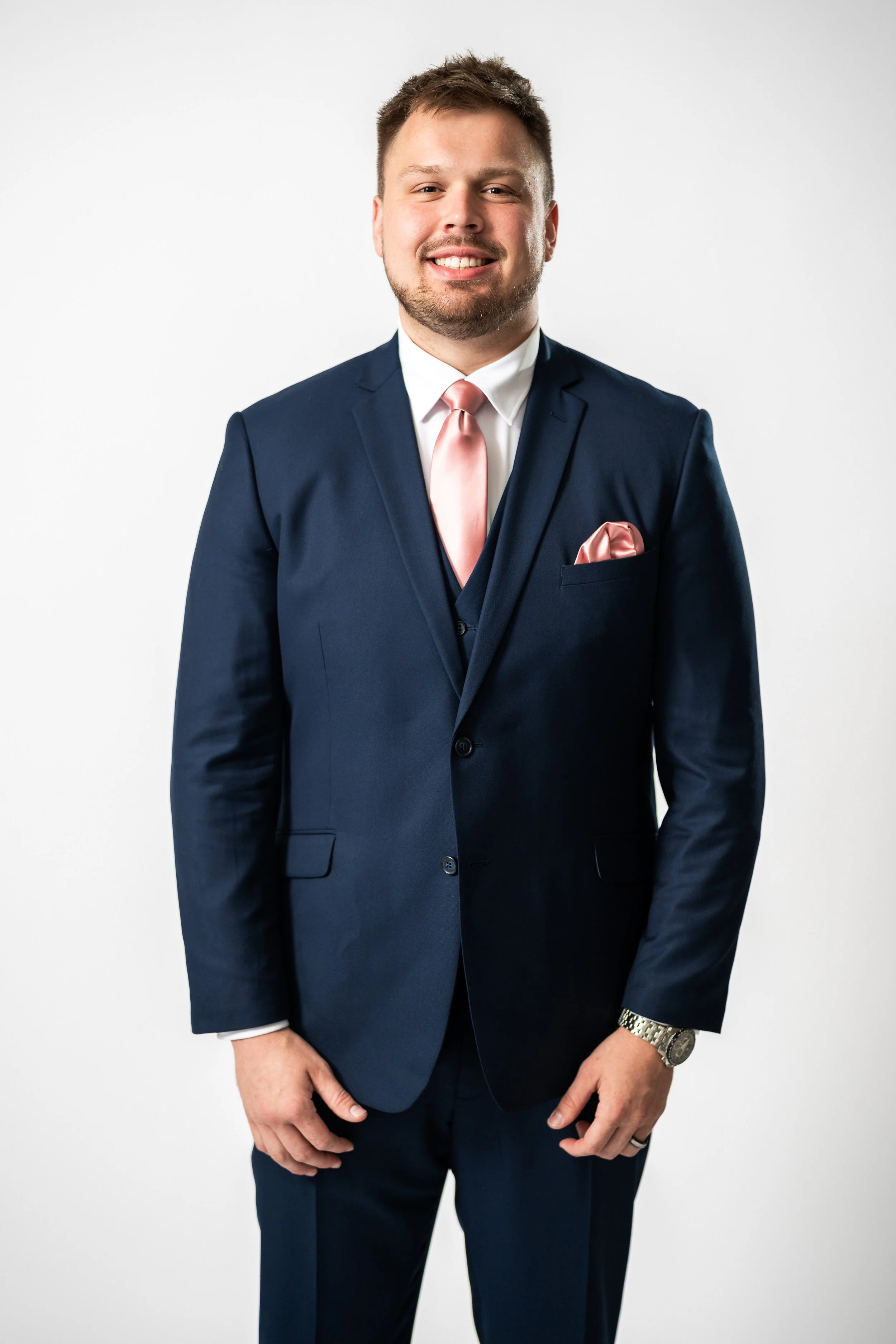 A man wearing a navy blue suit with a pink tie and pocket square, standing against a plain white background.