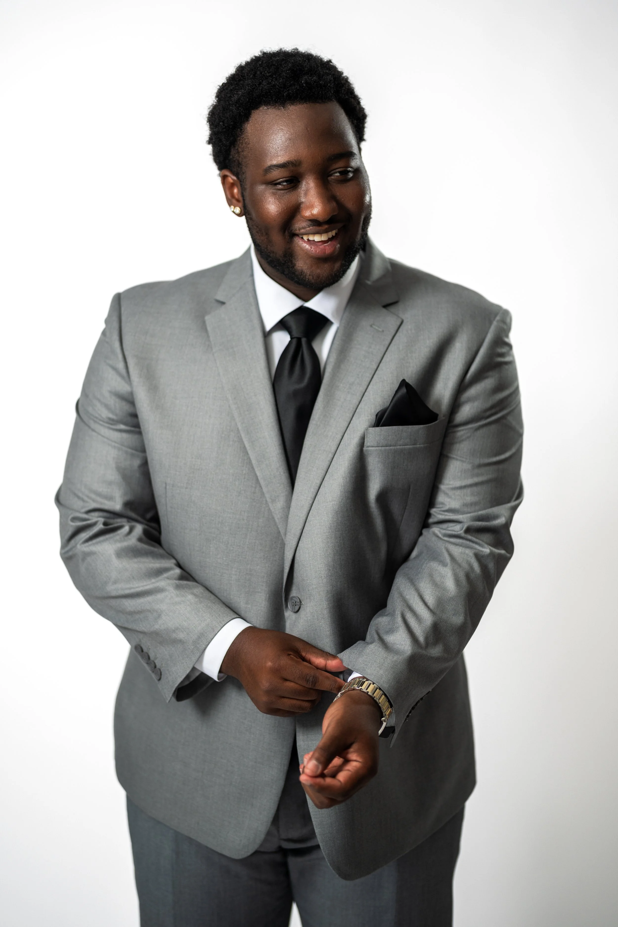 A young man in a gray suit, white shirt, and black tie adjusting his watch, standing against a plain white background.