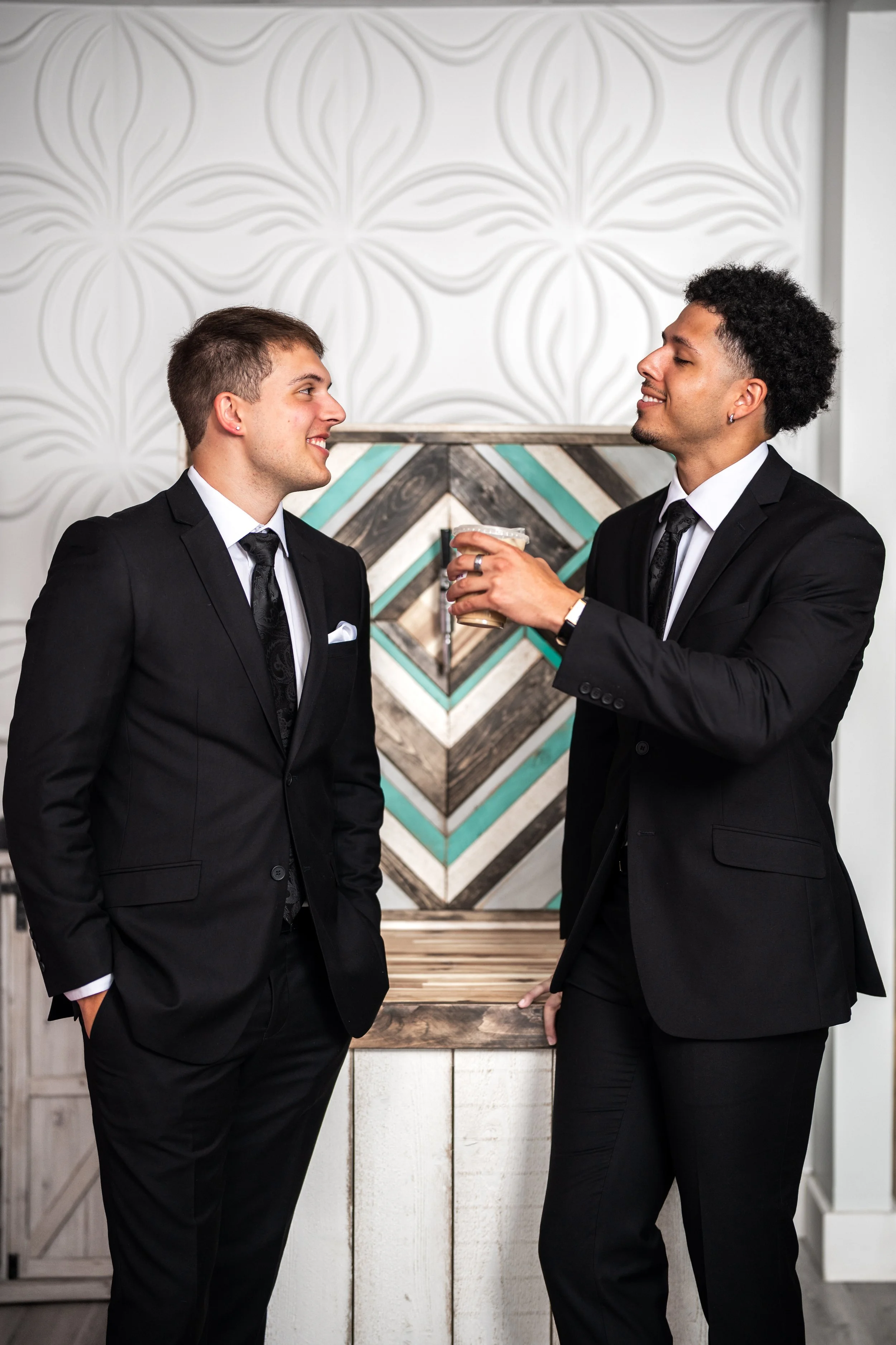 Two men dressed in black suits talking and smiling, one holding a drink, standing in a modern interior with a textured wall and a geometric artwork in the background.