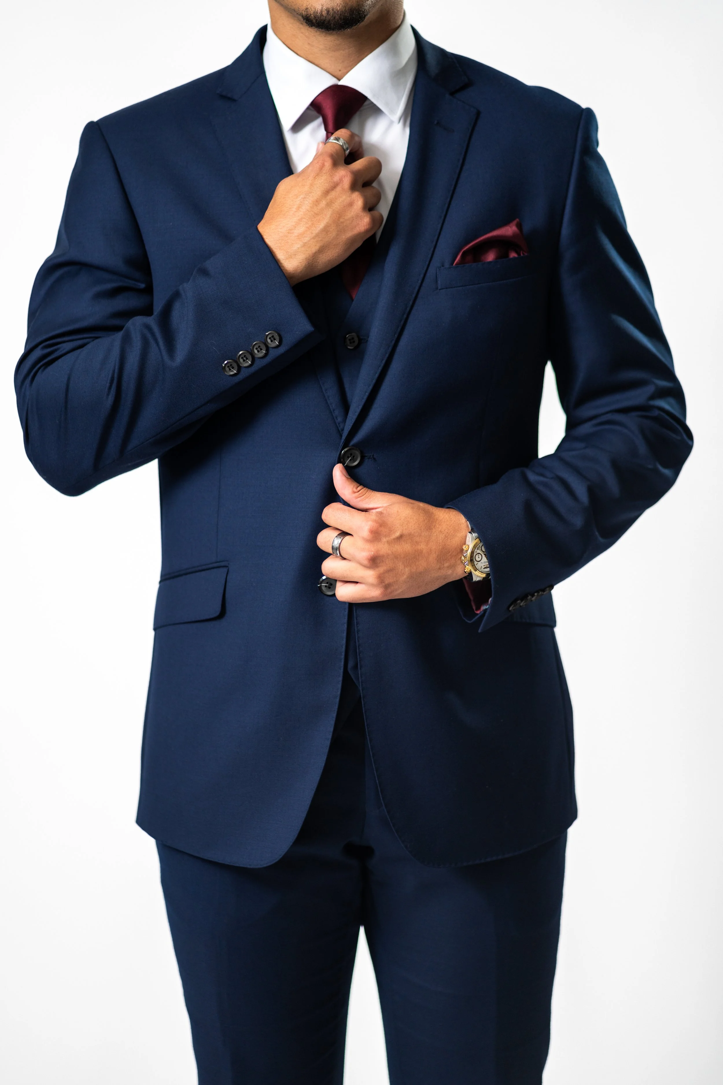 A man wearing a navy blue suit with a white dress shirt, maroon tie, and matching pocket square, adjusting his suit jacket.