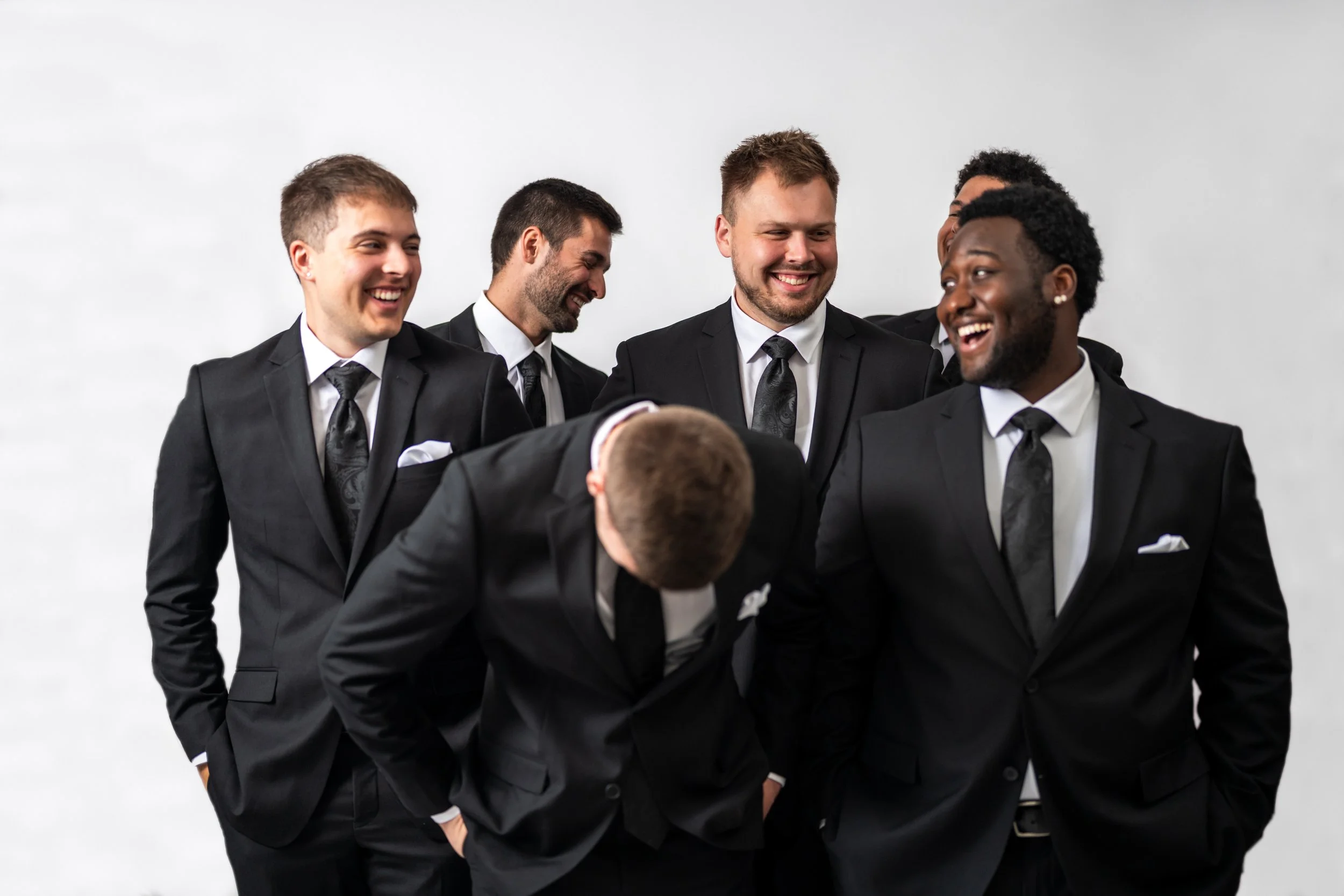 Group of six men in black suits and ties laughing and smiling together in front of a plain white background.