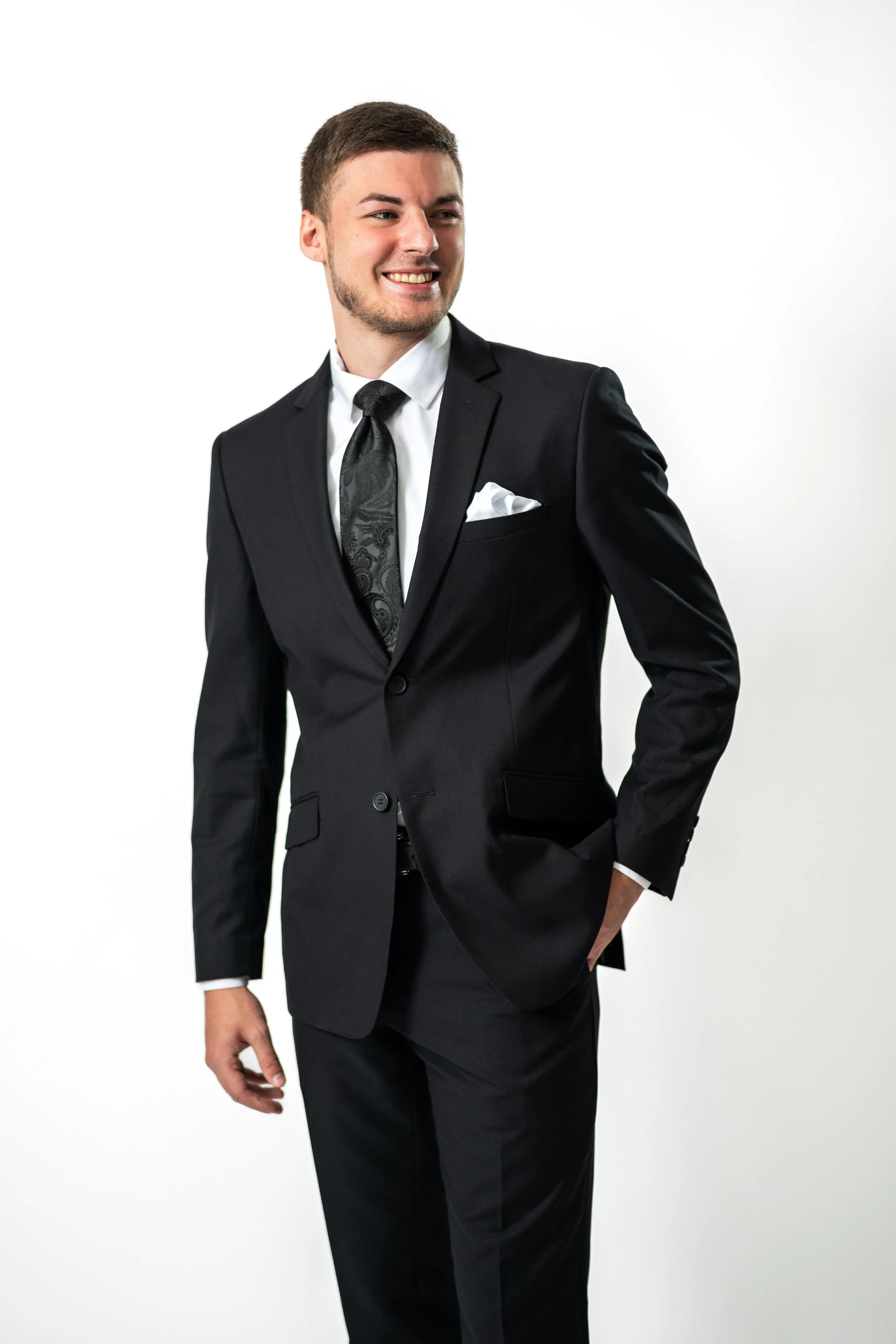 A young man in a black suit, white shirt, and patterned black tie, smiling and standing with one hand in his pocket.