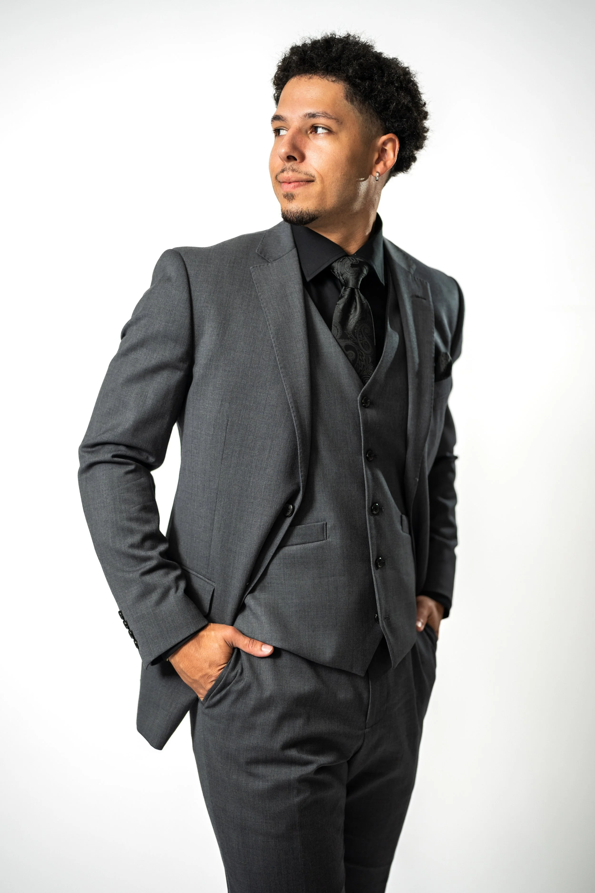 A man in a gray suit with a black shirt and tie, standing confidently with one hand in his pocket, looking to the side against a plain white background.