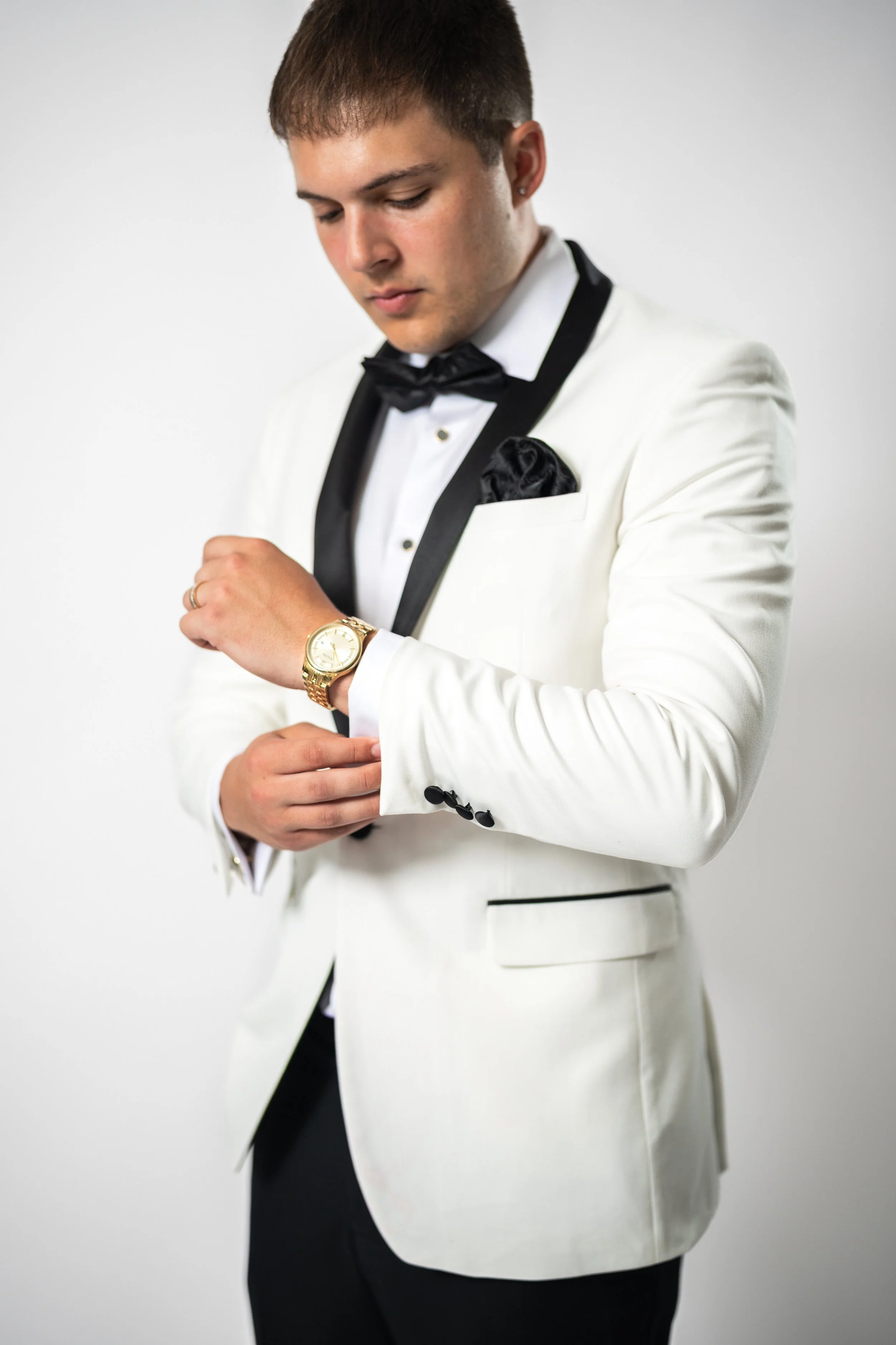 A young man in a white tuxedo with black accents, wearing a black bow tie, black dress pants, and a black pocket square, looks down at his gold wristwatch.