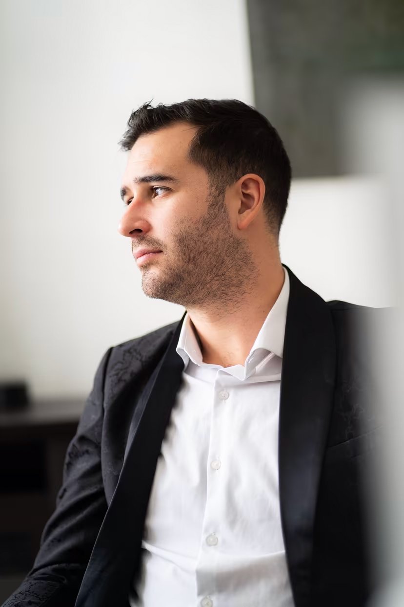 Homme en chemise blanche et veste noire, regardant pensivement vers la gauche dans un environnement intérieur éclairé.
