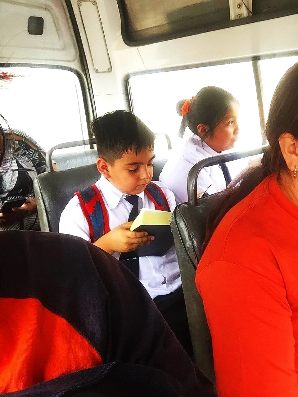 Niño en uniforme escolar leyendo un libro en un autobús, acompañado de otras personas, con ventanas al fondo.