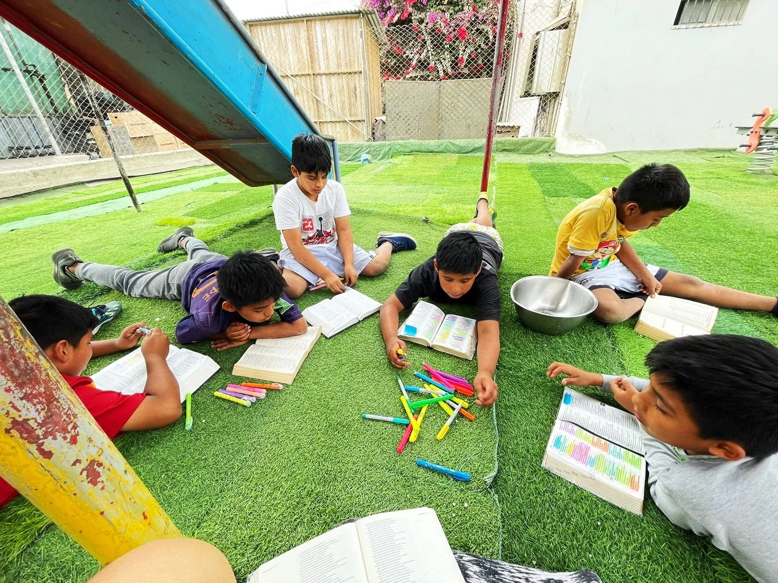 Niños leyendo libros mientras están tumbados en césped artificial en un patio con resbaladilla y arena. Algunos niños tienen marcadores de colores y otros tienen un bol grande de metal.