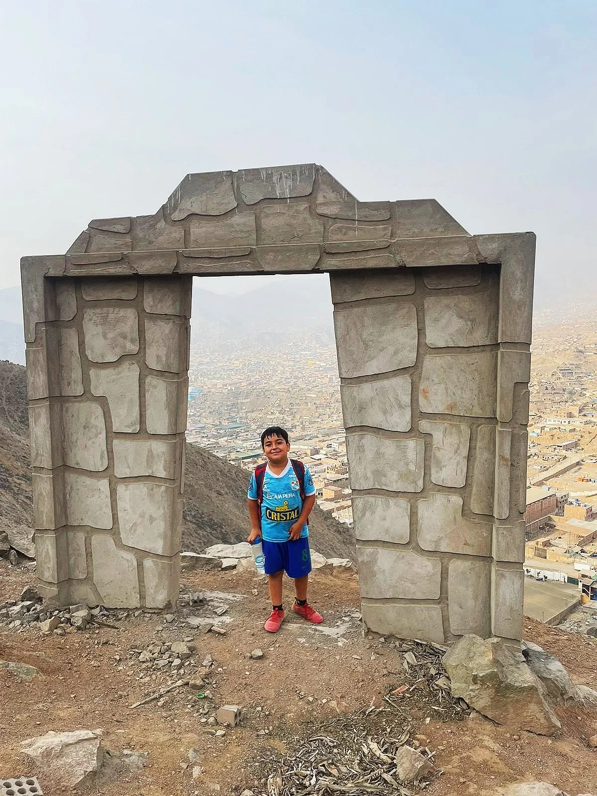 Niño sonriendo con mochila y botella de agua, de pie frente a una estructura de piedra en una colina con vista de la ciudad en el fondo.
