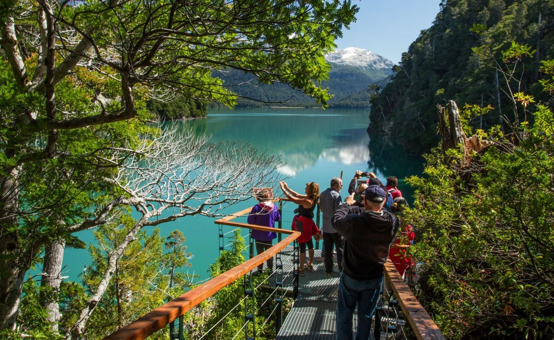 Tourists on a viewing platform overlooking a turquoise lake surrounded by green trees and mountains, with some snow-capped peaks in the distance.