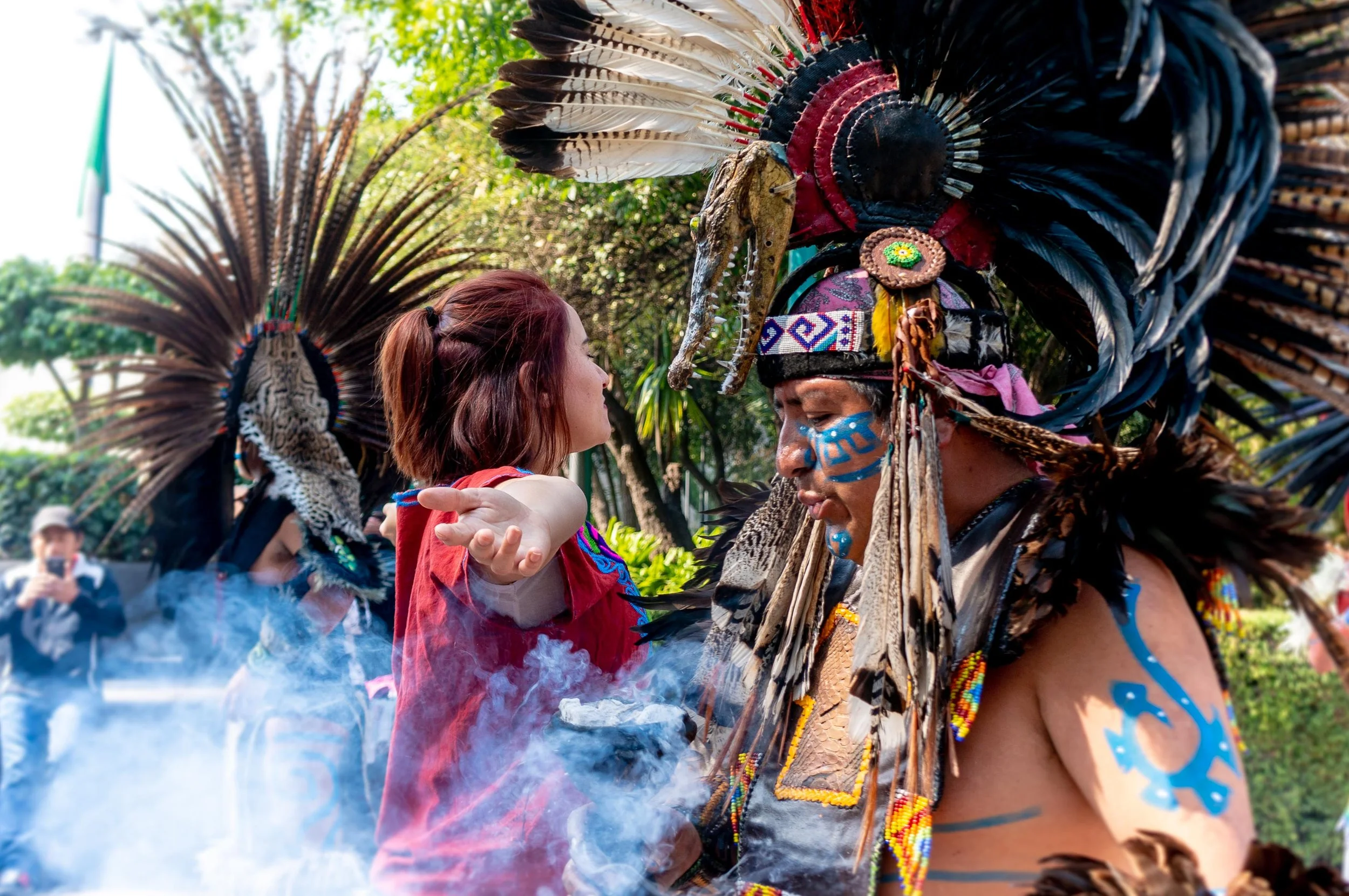 A person dressed in traditional indigenous attire with a large feather headdress faces a woman with short brown hair, who is reaching out with her arm. The scene appears to be part of a cultural event or celebration outdoors.