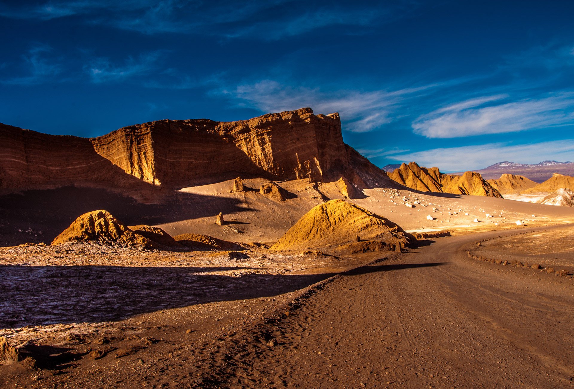 A dirt road through a desert landscape with layered red and orange rock formations and mountains in the distance, under a partly cloudy blue sky, Argentina.