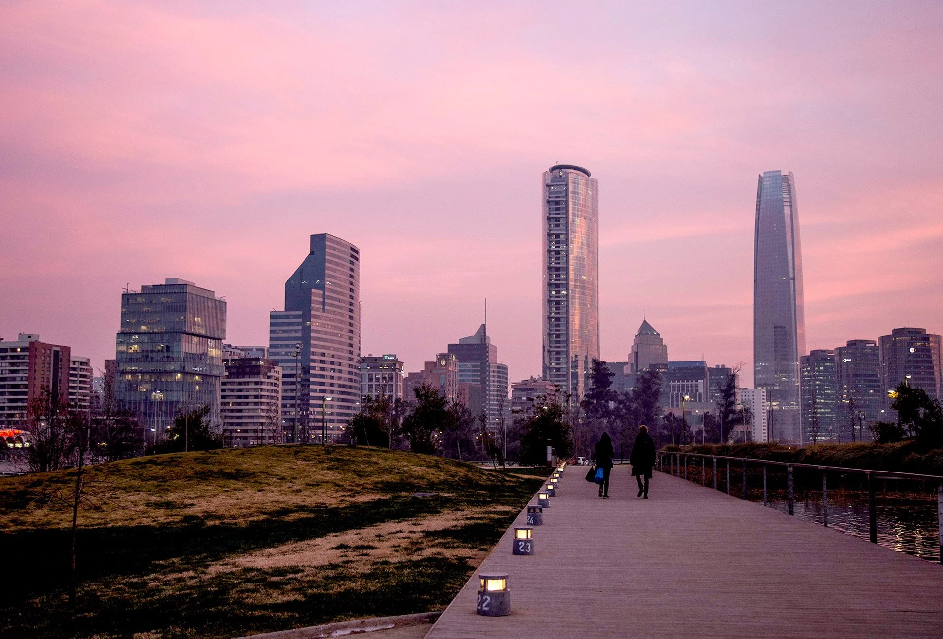 A city skyline during sunset with a pink and purple sky, tall modern buildings, and a walkway with two people walking along a river.