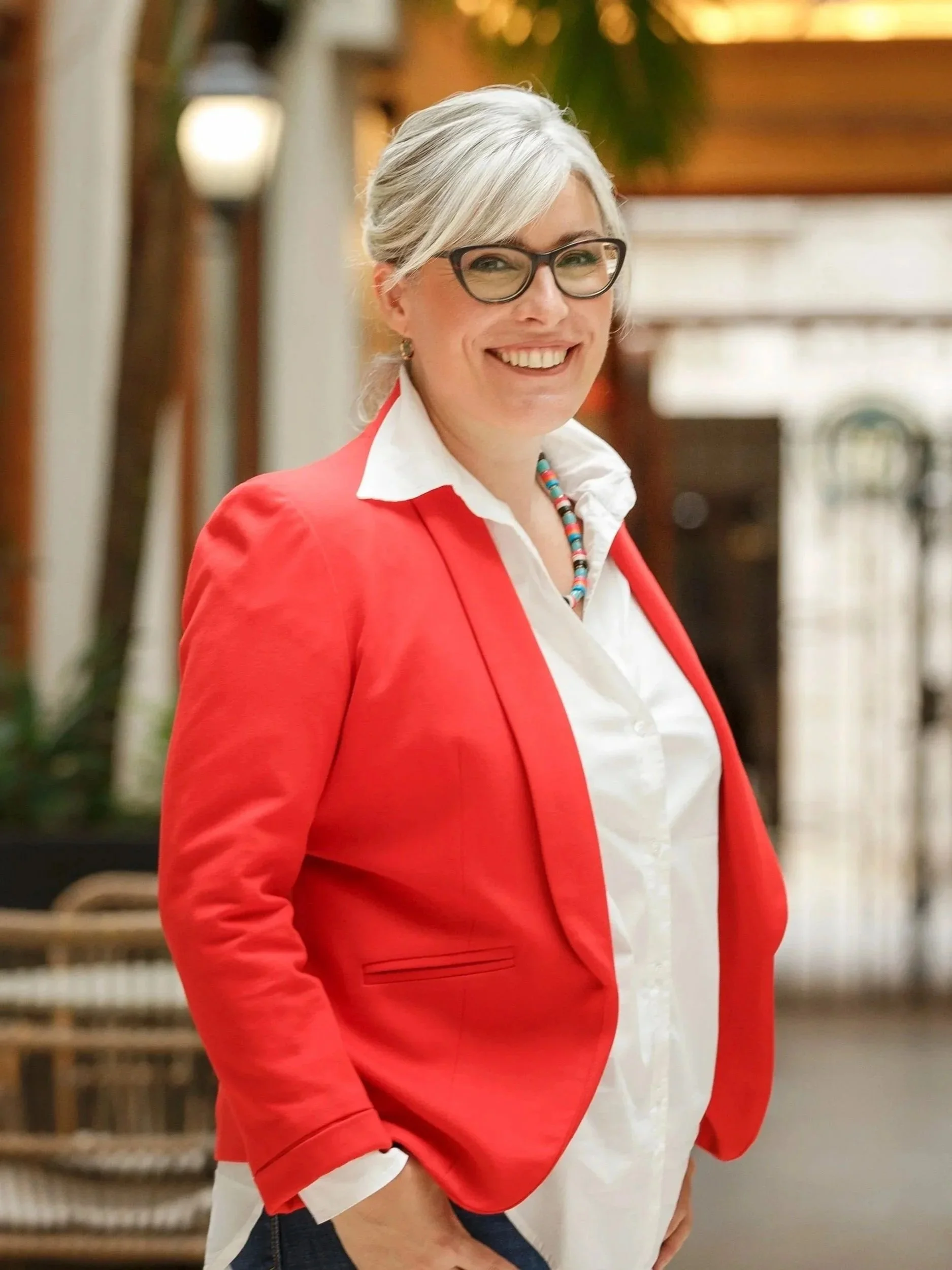 A woman with gray hair and glasses smiling outdoors, wearing a red blazer over a white shirt and a colourful beaded necklace, Buenos Aires Argentina.